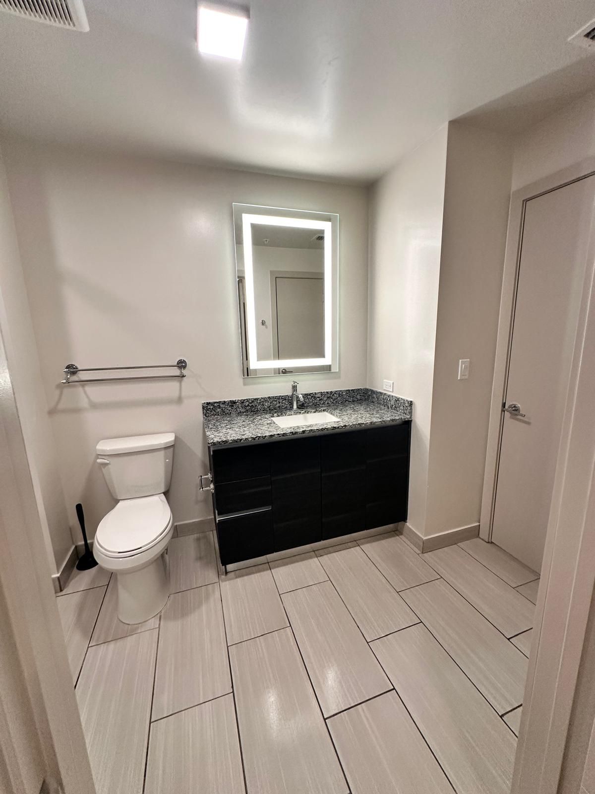 Bathroom with a toilet, vanity with lighted mirror, and tile flooring.