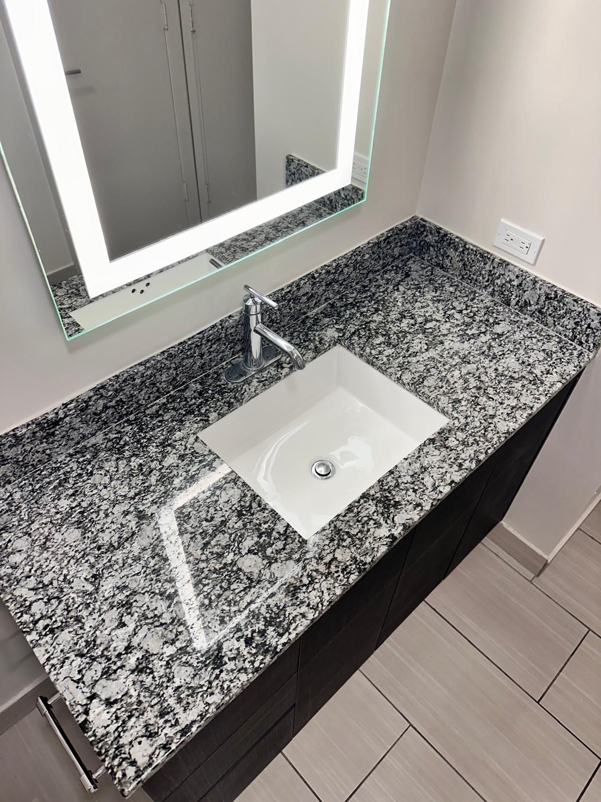 Vanity with granite countertop, square sink, lit mirror, chrome faucet, and electrical outlet.