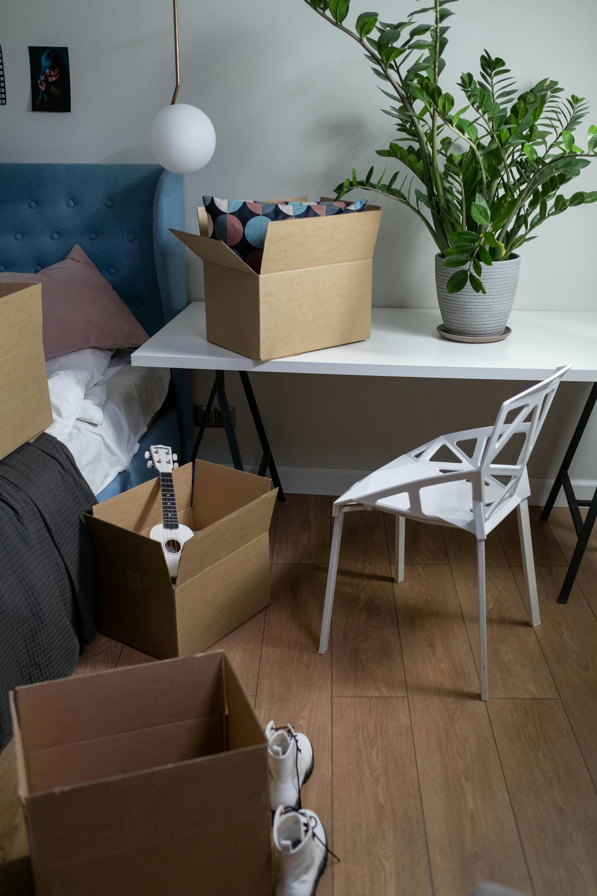 Cardboard boxes and a guitar on a wooden floor, next to a desk with a plant and a white chair.