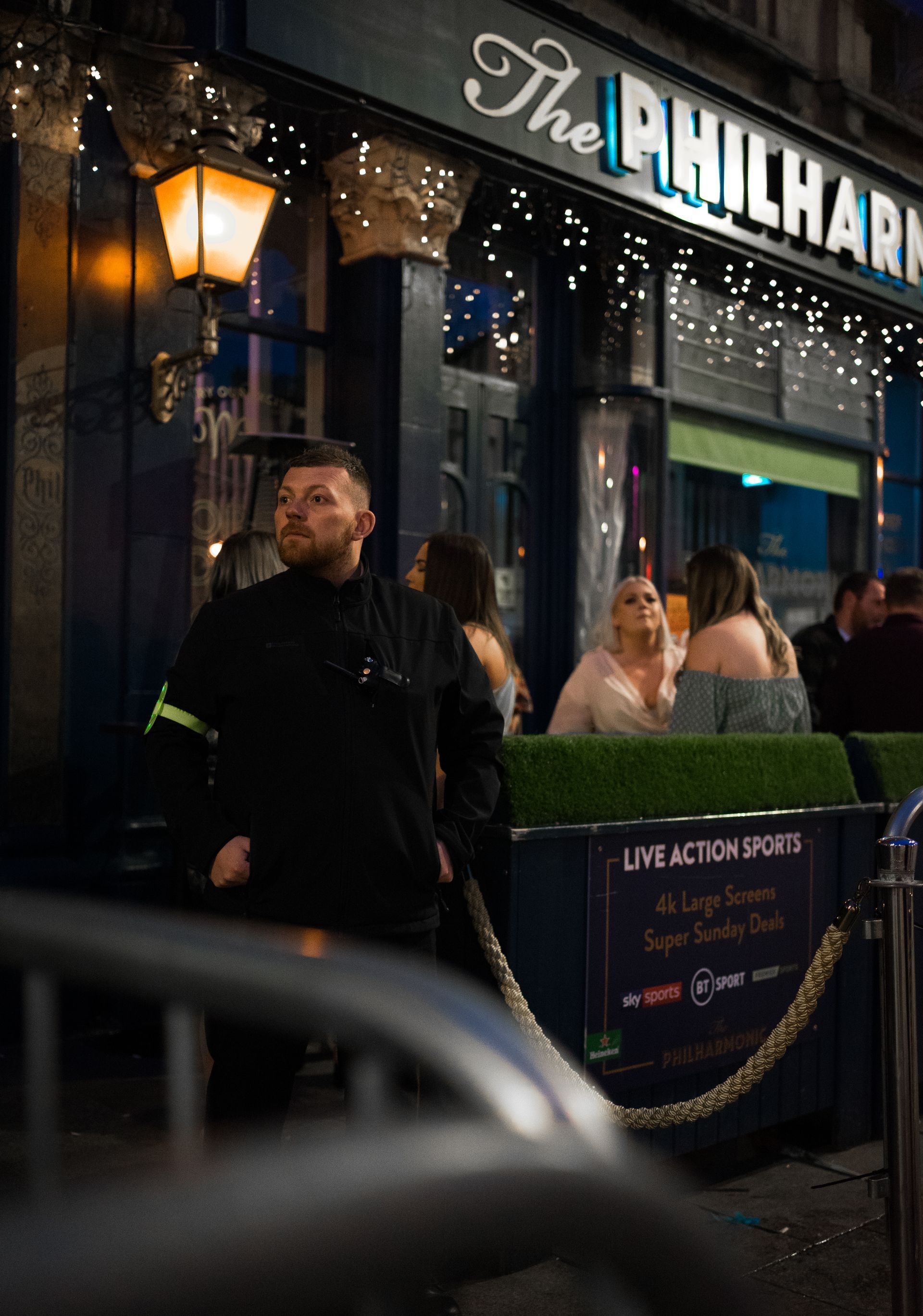 A man in black uniform stands outside The Philharmonic pub, people seated at outdoor tables.