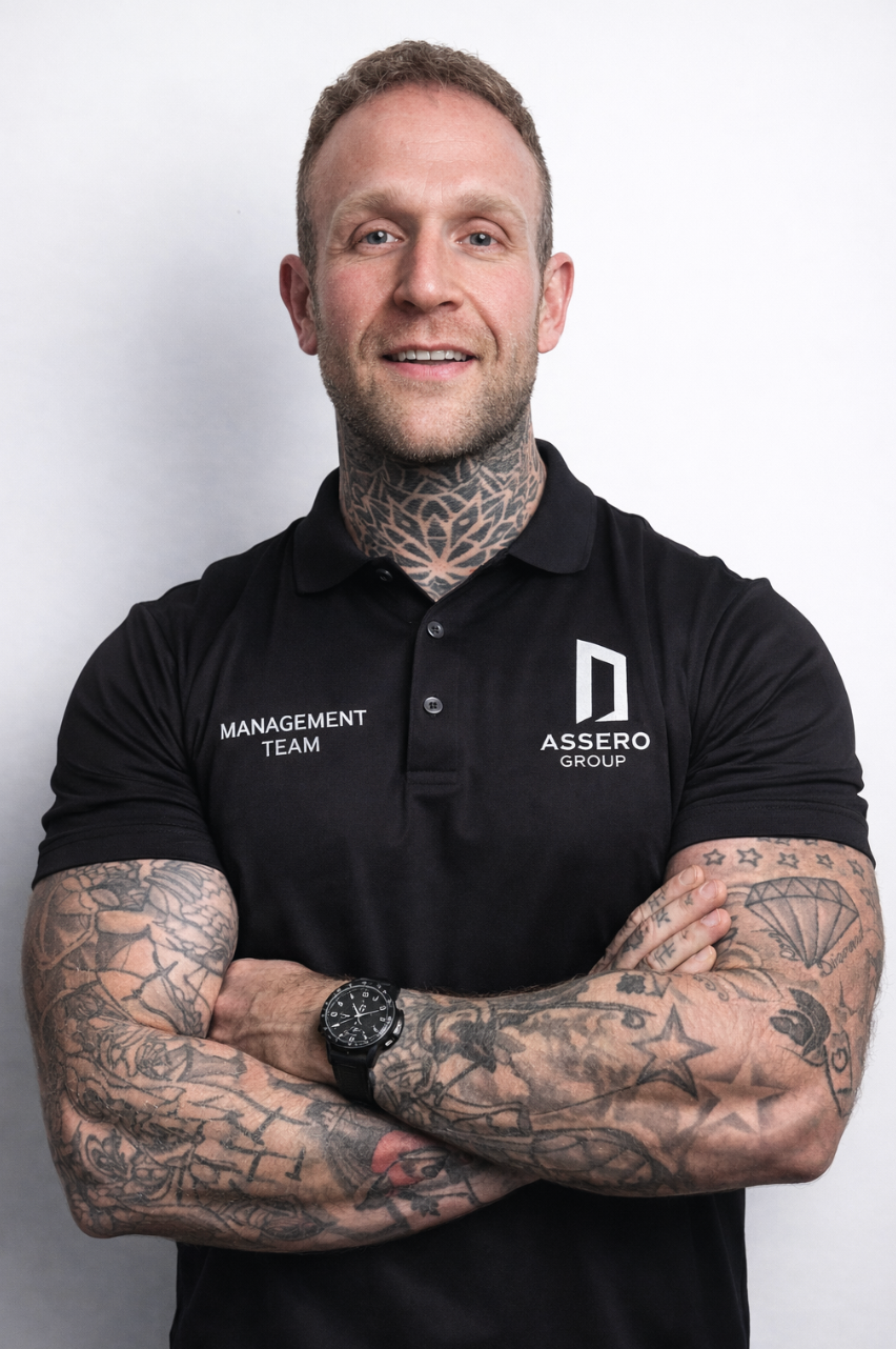 A person with tattoos on their neck and arms wearing a black polo shirt with a logo, posing with their arms crossed.