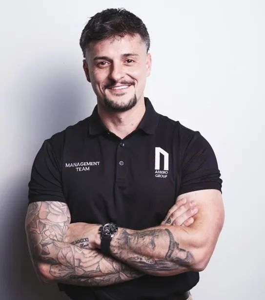 A smiling person wearing a black polo with an embroidered logo, arms crossed, displaying arm tattoos against a white wall.