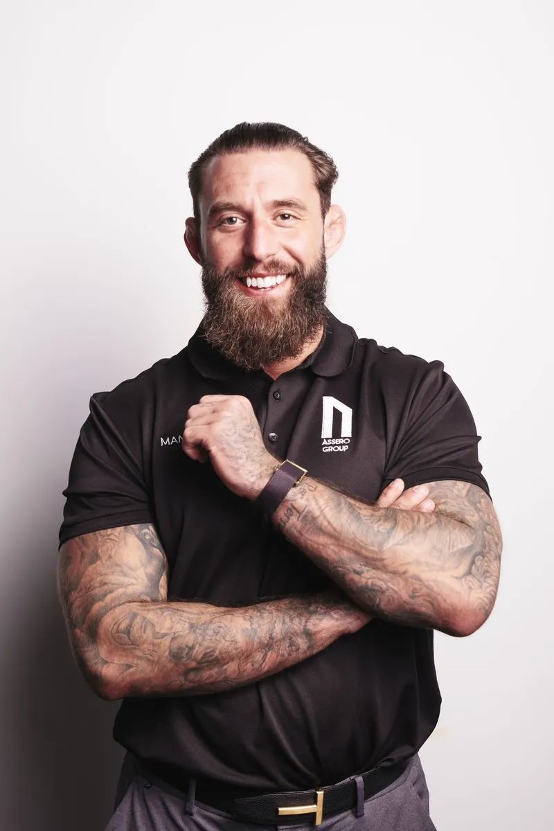 A bearded person in a black polo with a logo, smiling with arms crossed against a white background.