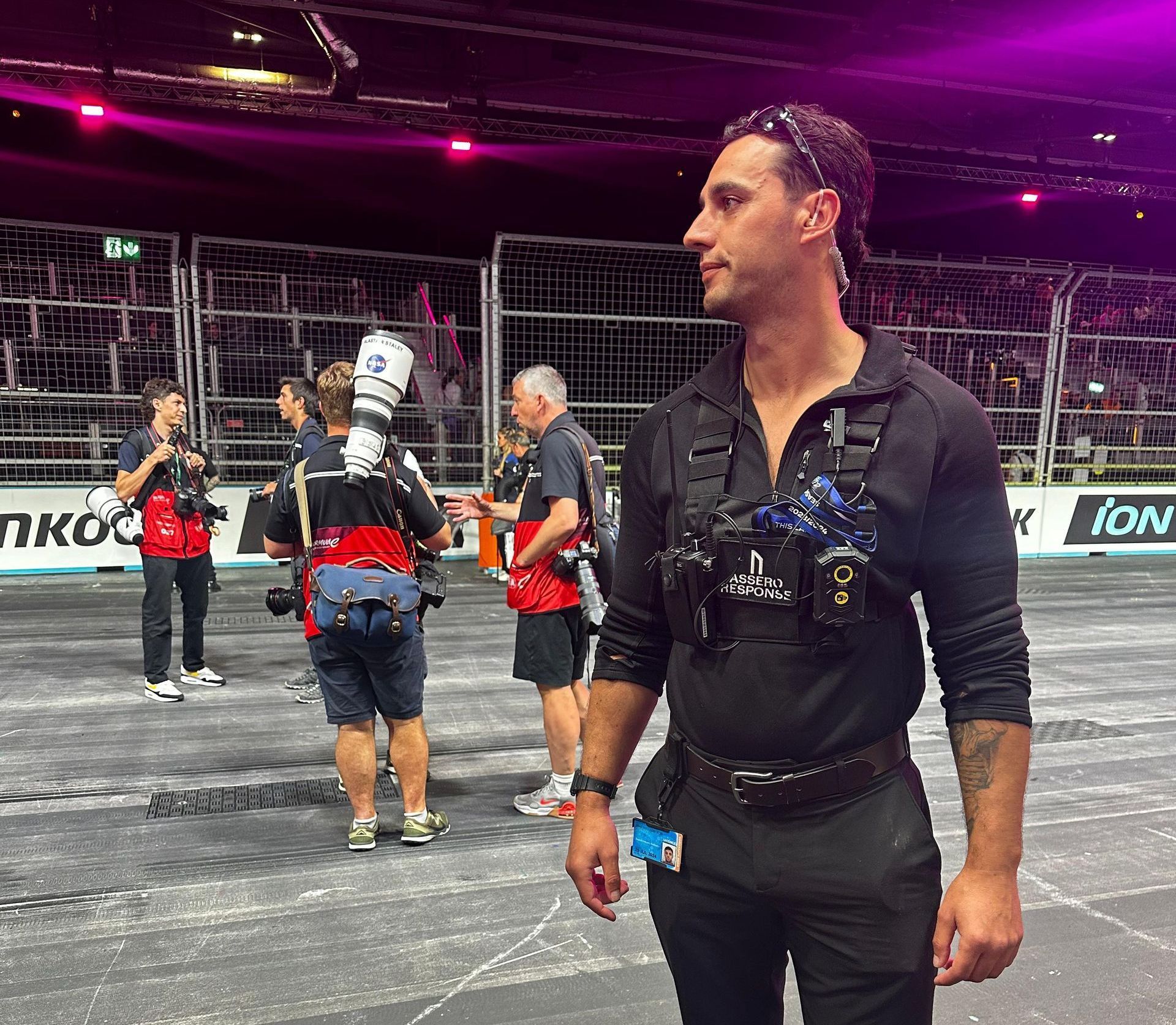 Man wearing a chest rig and looking off-camera. Other people with cameras stand nearby on a race track.