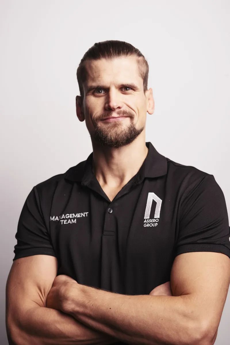 A man with a short beard and hair tied back, wearing a black polo shirt, posing with arms crossed against a white wall.
