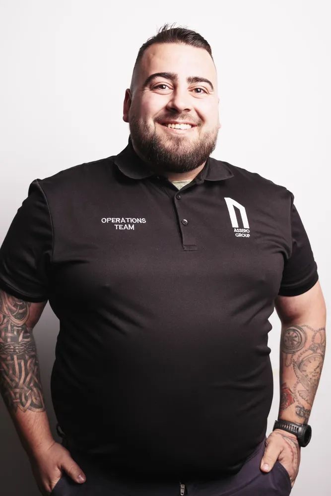 A smiling person with arm tattoos wearing a black polo shirt labeled