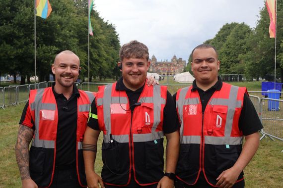 Three people wearing matching black shirts and red high-visibility vests smile while standing outdoors at an event.