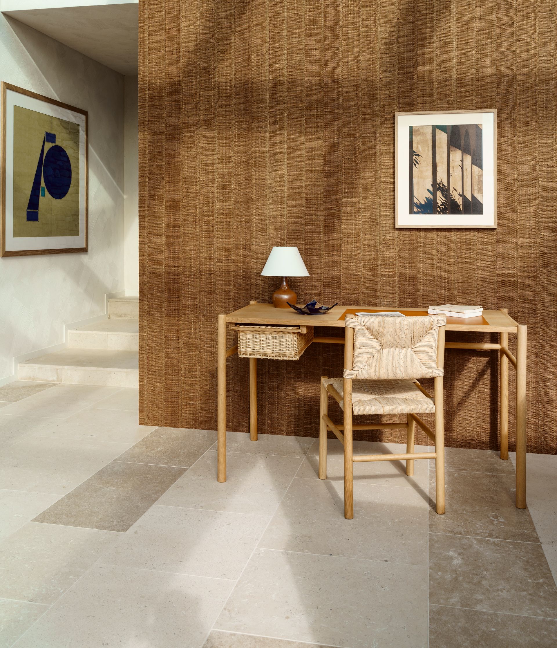 Scrivania con lampada e sedia contro una parete marrone testurizzata, opere d'arte sulla parete laterale, pavimento neutro, interni luminosi.