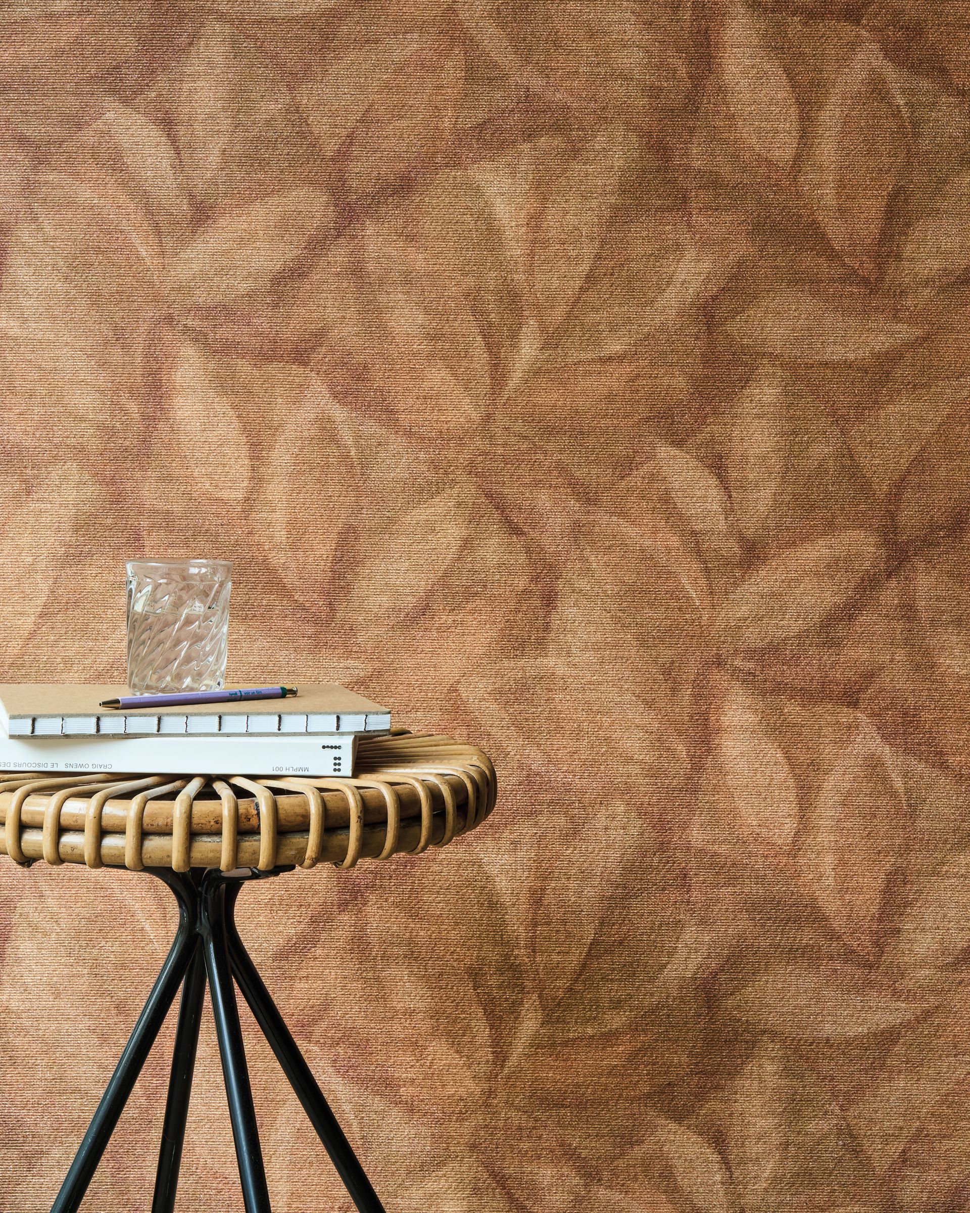 Carta da parati testurizzata marrone con un piccolo tavolo in vimini su cui sono appoggiati libri e un bicchiere.