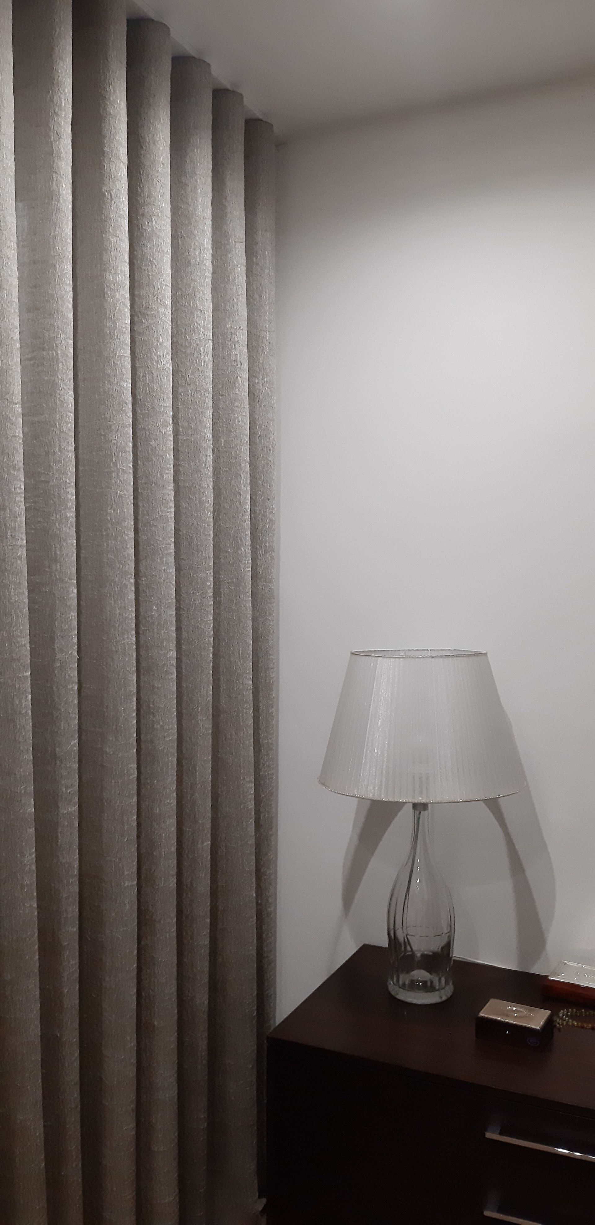 Tende beige accanto a una parete bianca, comodino scuro con lampada sopra.