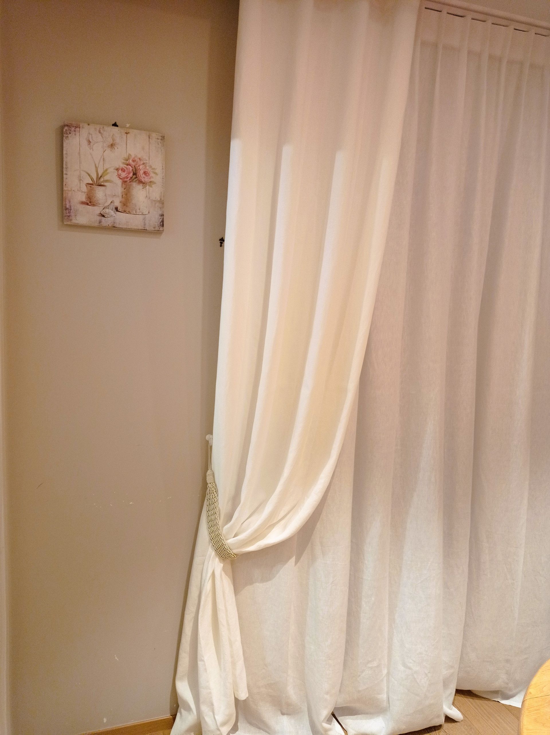 Tende color crema drappeggiate e legate, accanto a una parete chiara con un piccolo dipinto floreale.