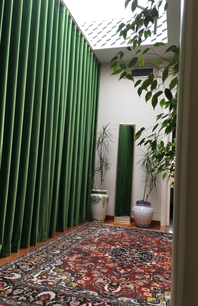 Camera con tende di velluto verde, tappeto decorato, piante in vaso e lucernario.