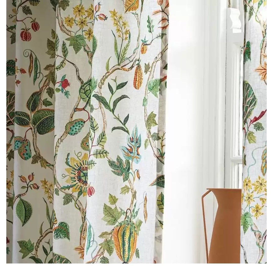 Tende con motivi floreali nei colori verde, giallo, arancione e marrone sono appese vicino a una finestra bianca e a una brocca marrone.