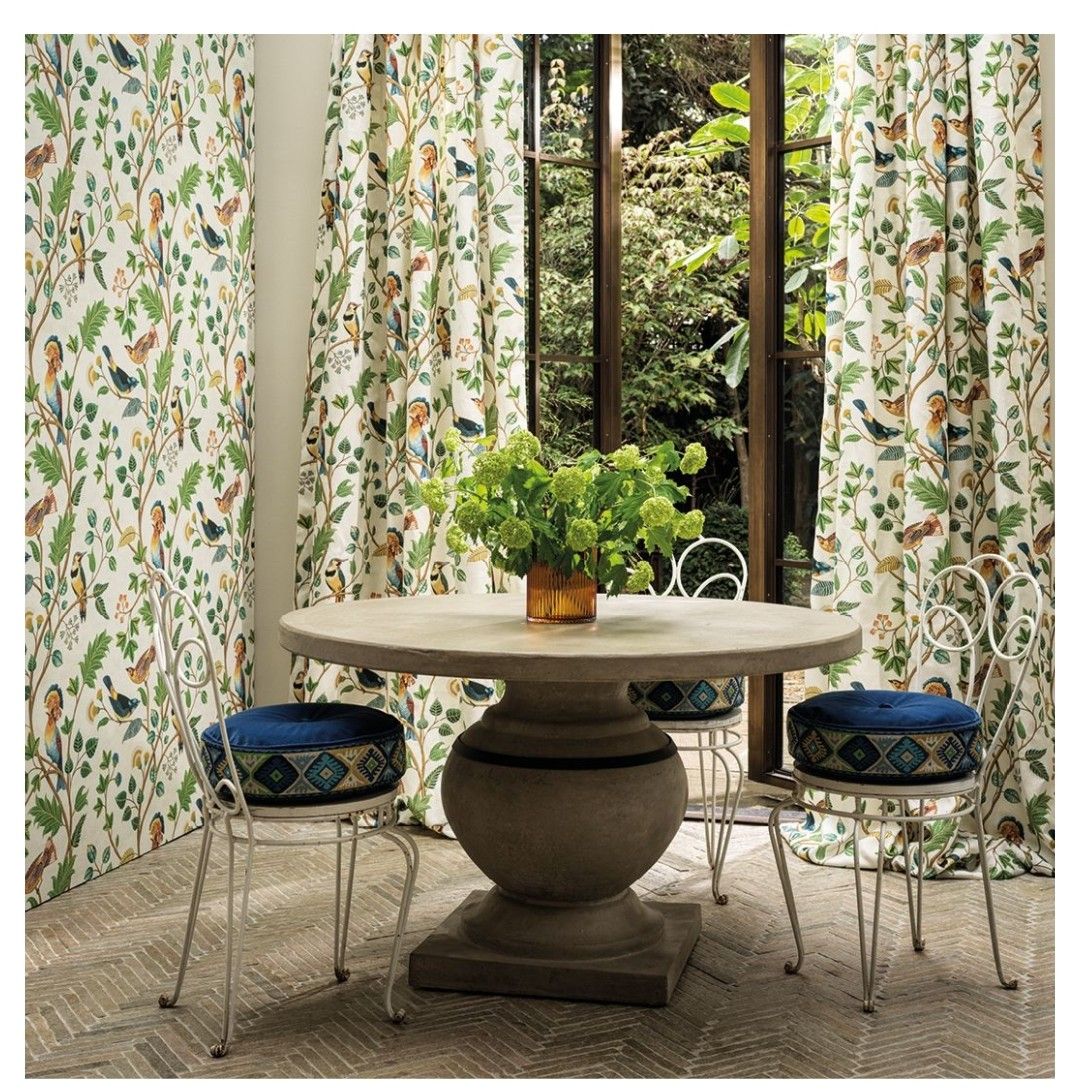 Interno con tavolo rotondo, tre sgabelli con cuscini blu e tende con motivi di uccelli e foglie.