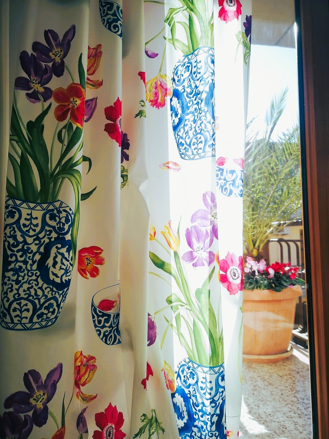 Tende bianche con vasi blu e fiori colorati incorniciano un balcone soleggiato.