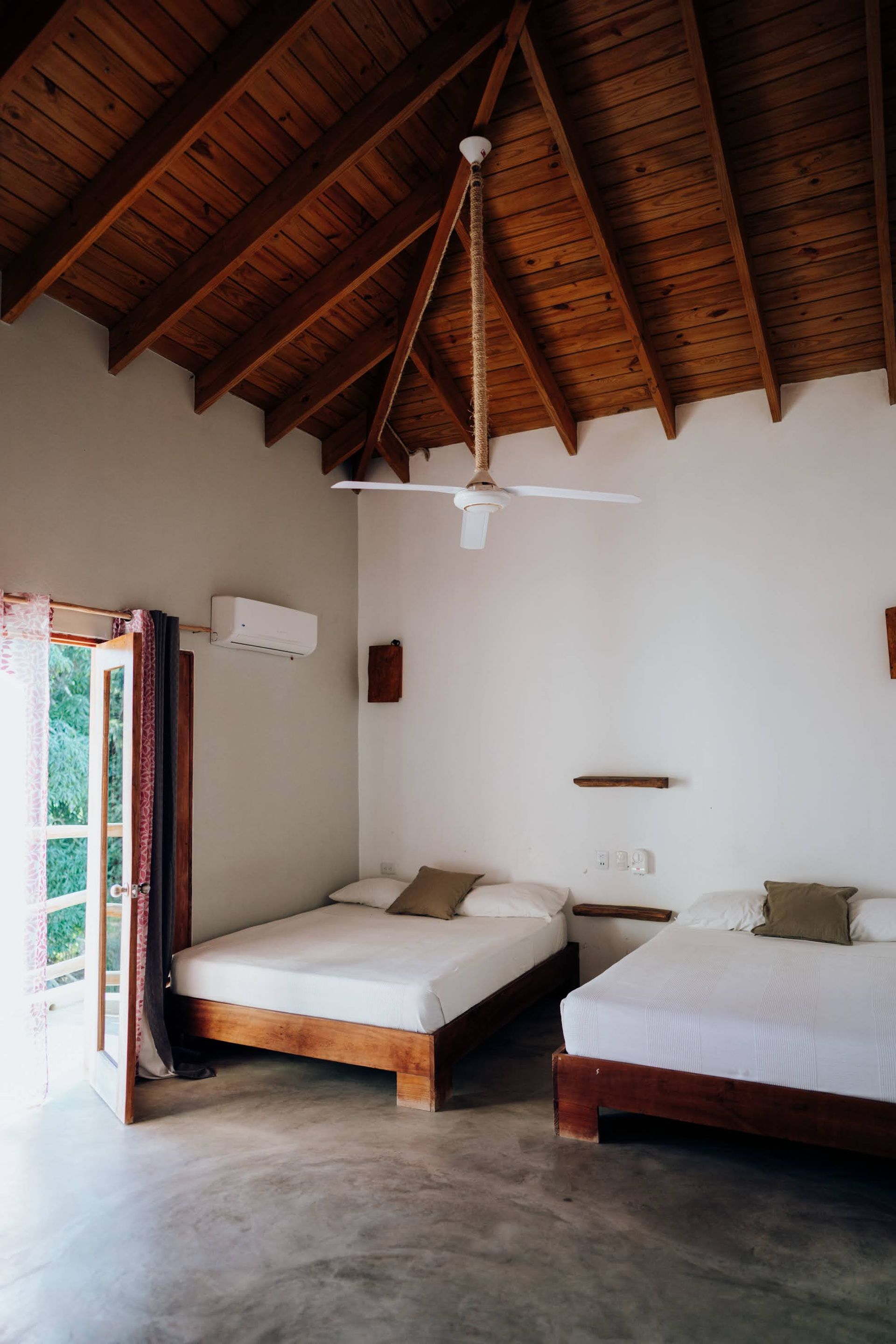 Bedroom with two beds, wooden ceiling, open door to greenery, white walls, ceiling fan.