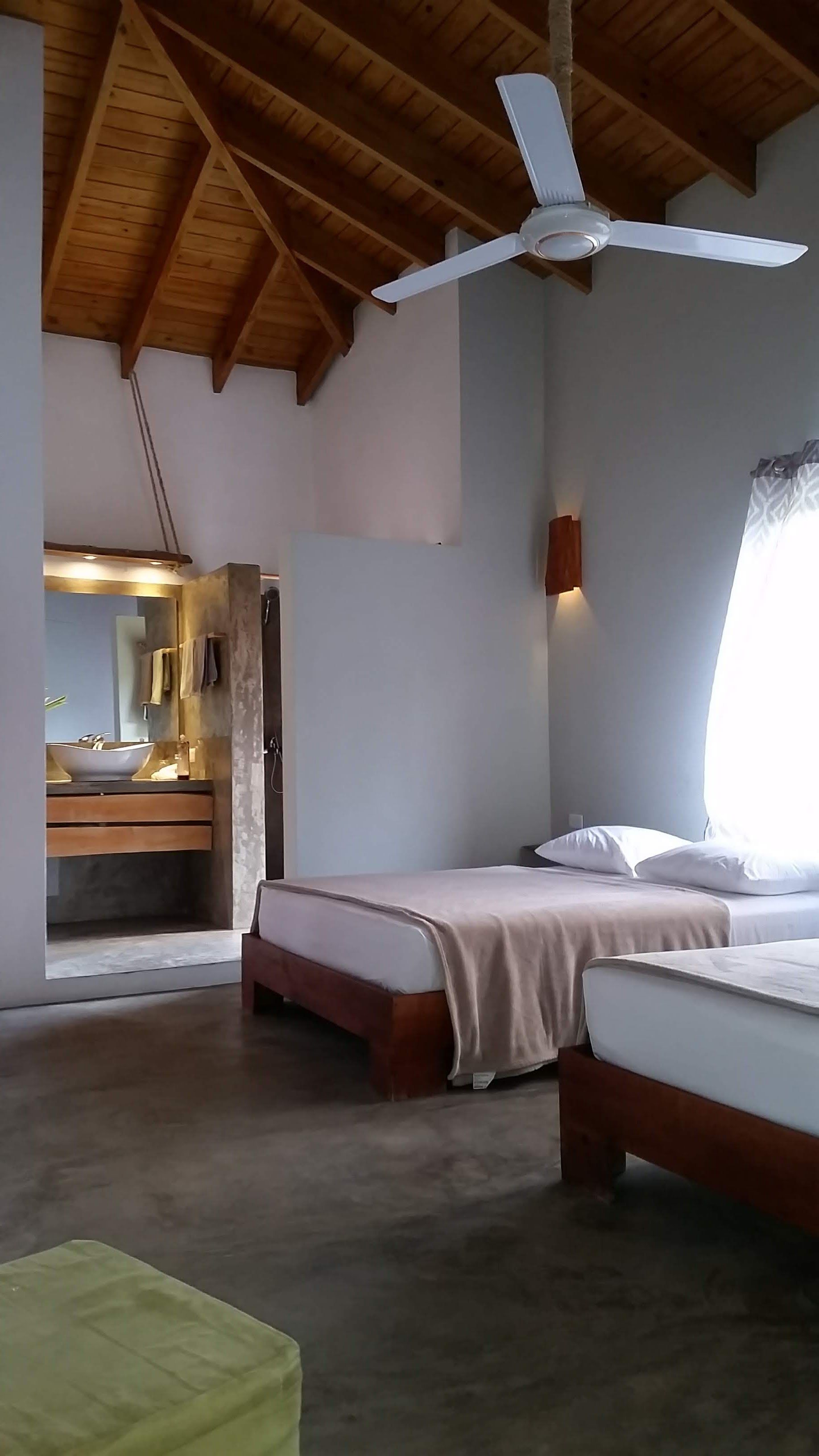 Bedroom with twin beds, bathroom visible, high wooden ceiling, white walls, and a ceiling fan.