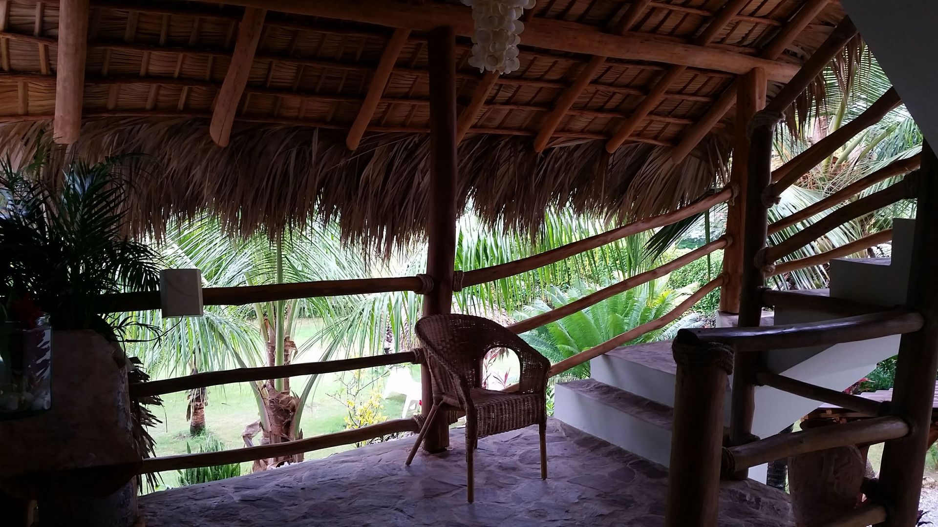 Rustic balcony with thatched roof, wooden railing, and a woven chair, overlooking a green landscape.