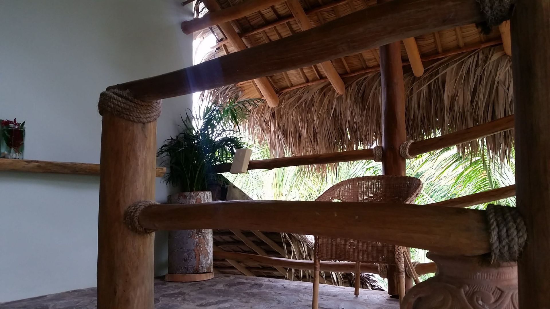 Wooden railing with rope accents, leading to a thatched roof area with wicker furniture and plants.