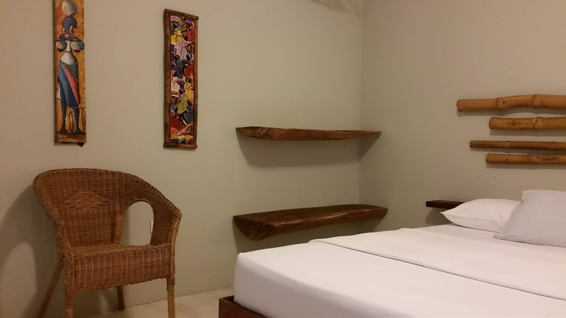 Bedroom with wicker chair, bed with white linens, wooden shelves, and wall art.