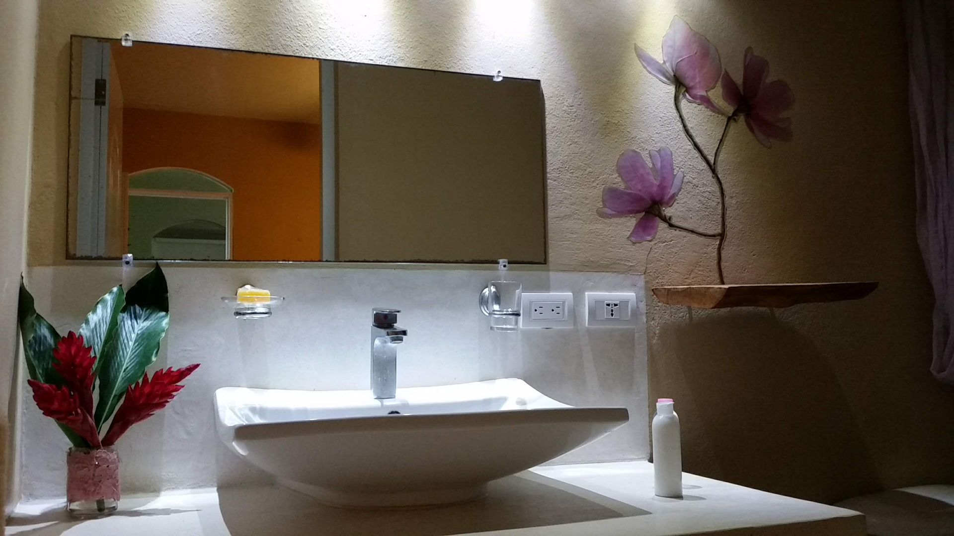 Bathroom sink with mirror, decorative flowers, and plants. Beige walls, white sink, and silver faucet.