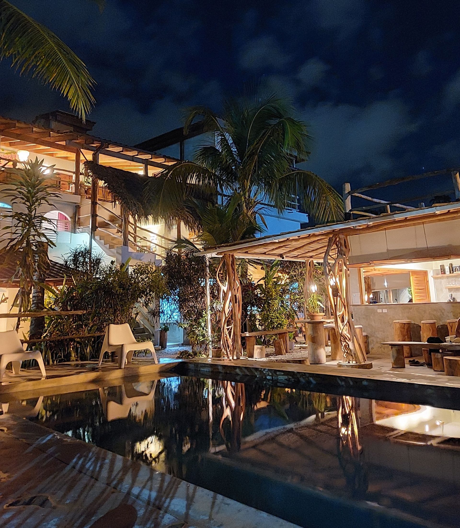 Nighttime view of a restaurant with a pool. The sky is dark, with lights reflecting on the water. Wooden structures and foliage are visible.