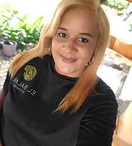 Blonde-haired person smiling, wearing a black shirt with logo. Outdoors with greenery in background.