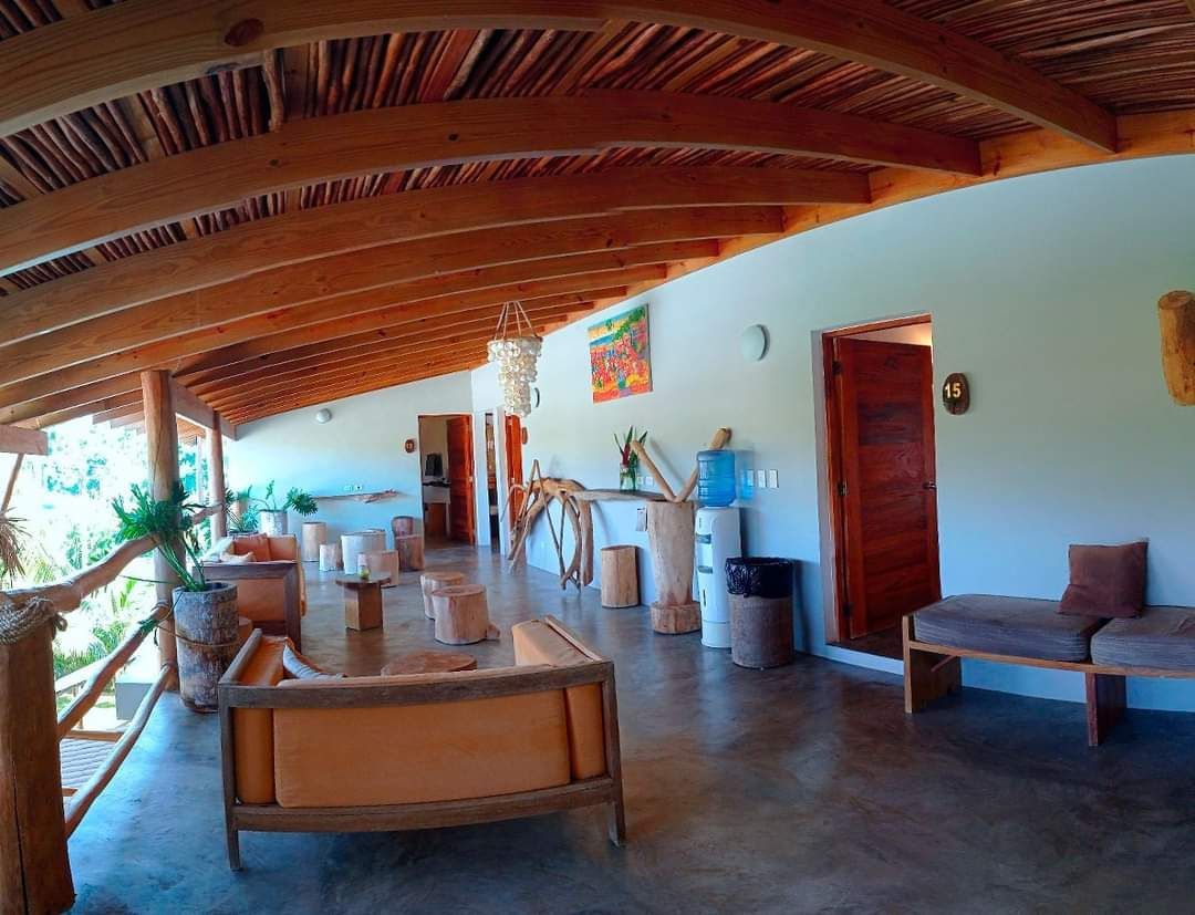 A rustic outdoor lounge with wooden beams, curved white walls, and seating, overlooking greenery.