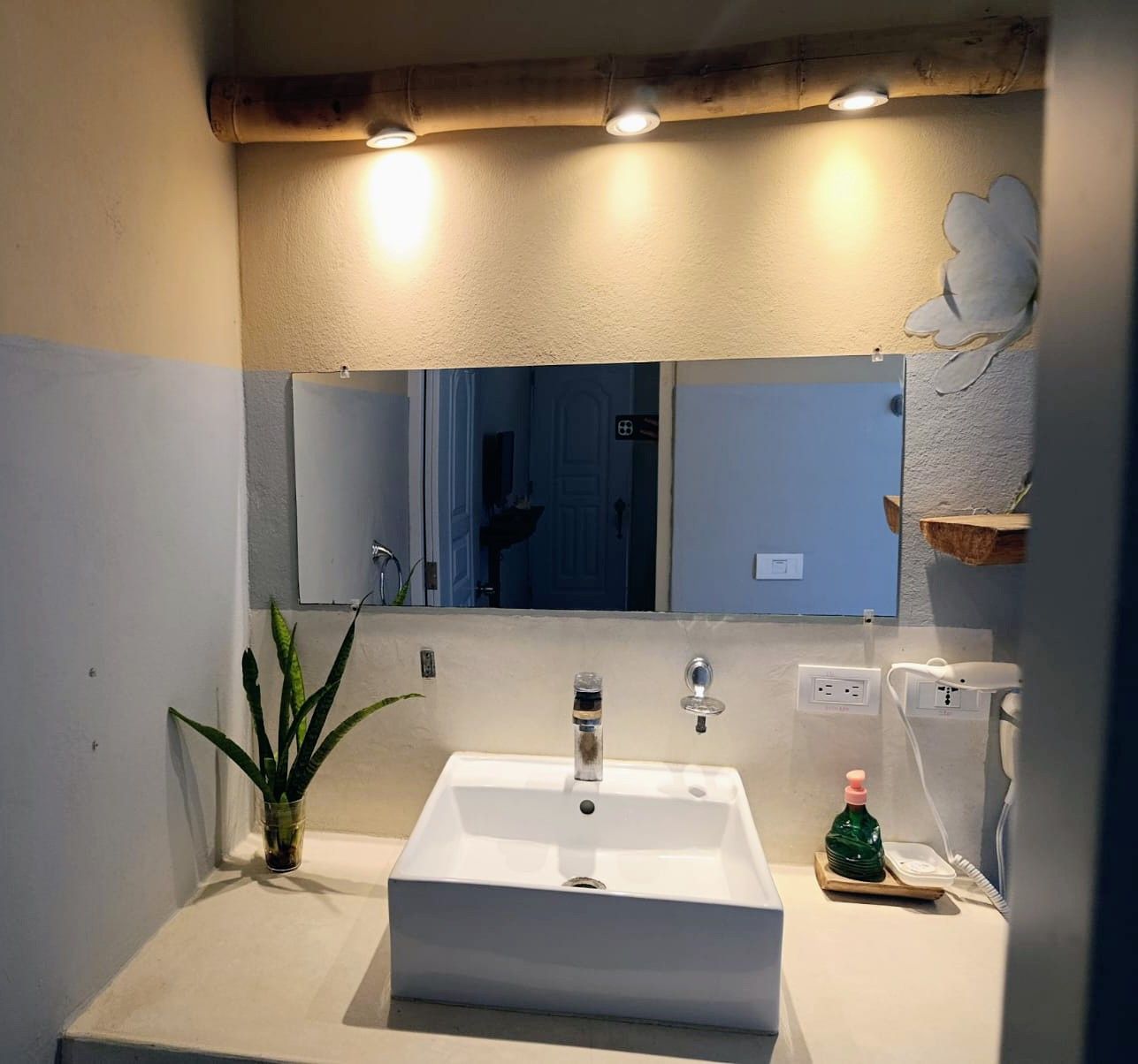 Bathroom with a white square sink, mirror, and a plant. Neutral walls with wood beam lighting.