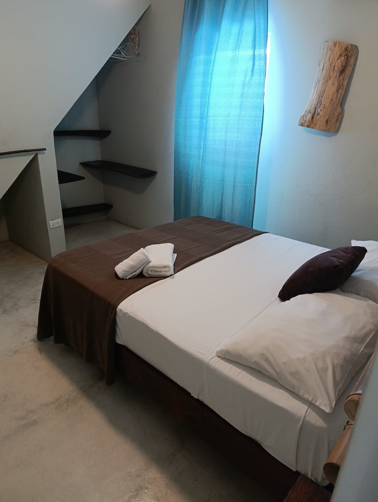 Bedroom with a bed covered in white linens, brown blanket, and blue curtains. Small wooden shelf on the wall.