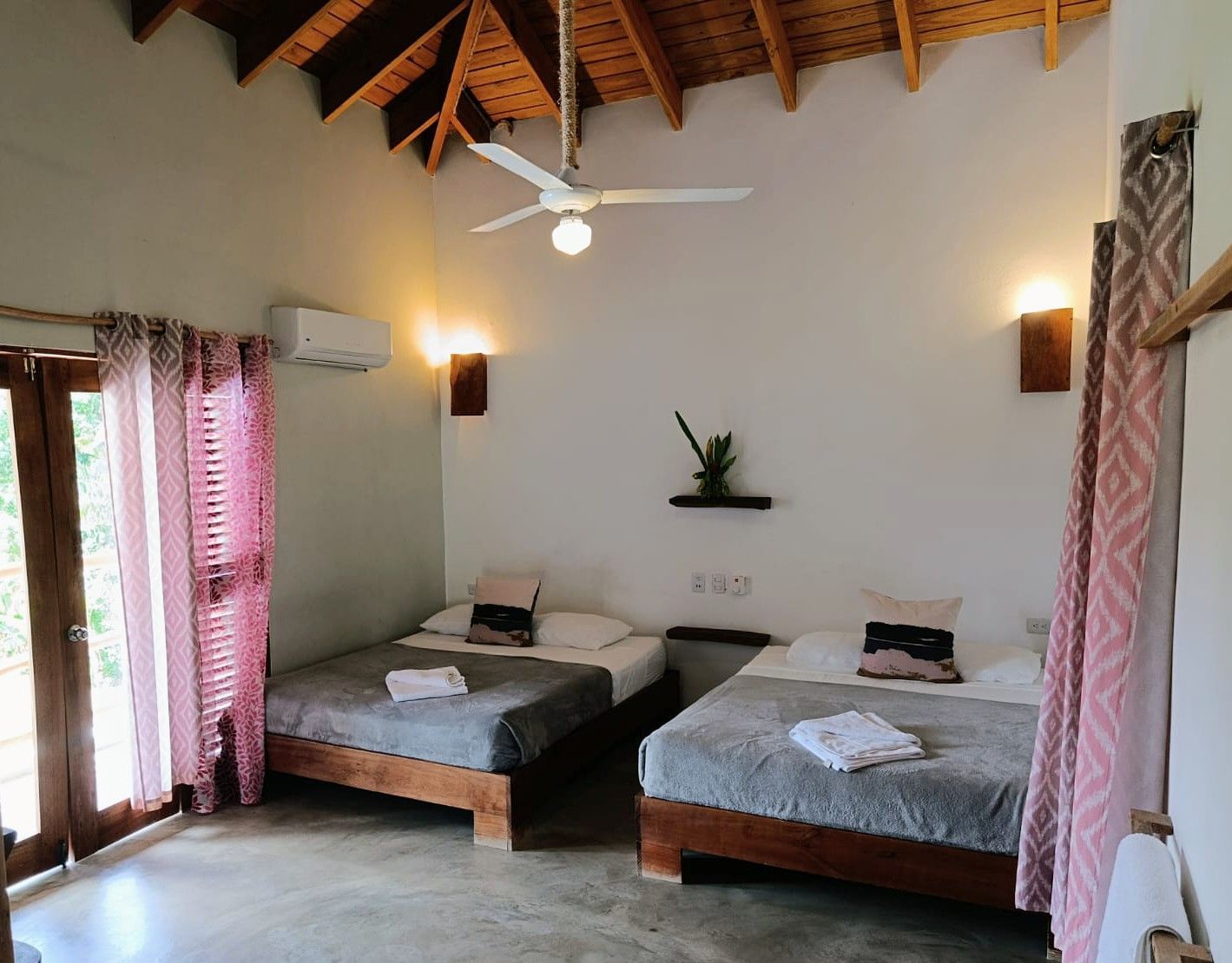 A bedroom with two beds, white walls, wooden ceiling, and a doorway with pink curtains.