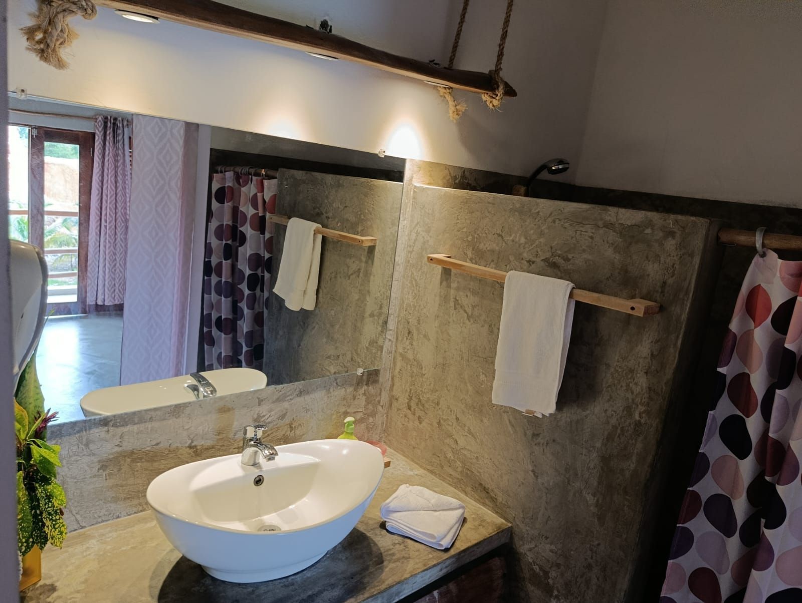 Bathroom with a white oval sink, mirror, concrete walls, towel racks, and patterned shower curtain.