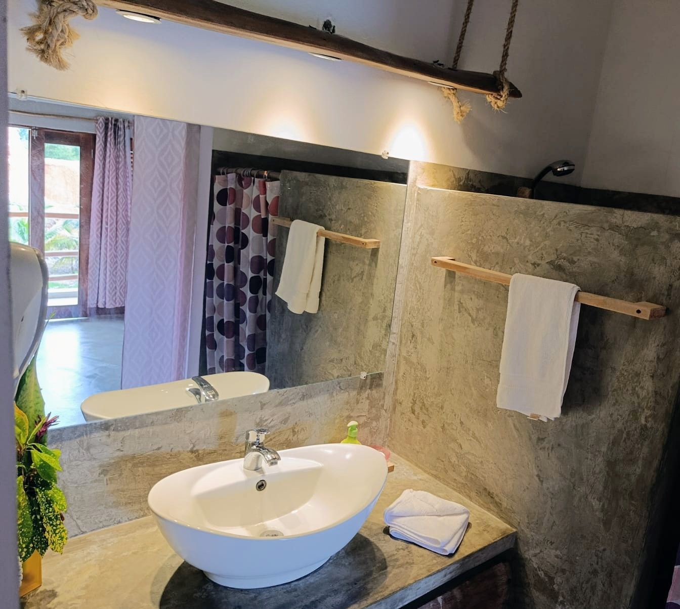 Bathroom with a concrete wall, a white oval sink, and a large mirror reflecting a bathtub and a doorway.