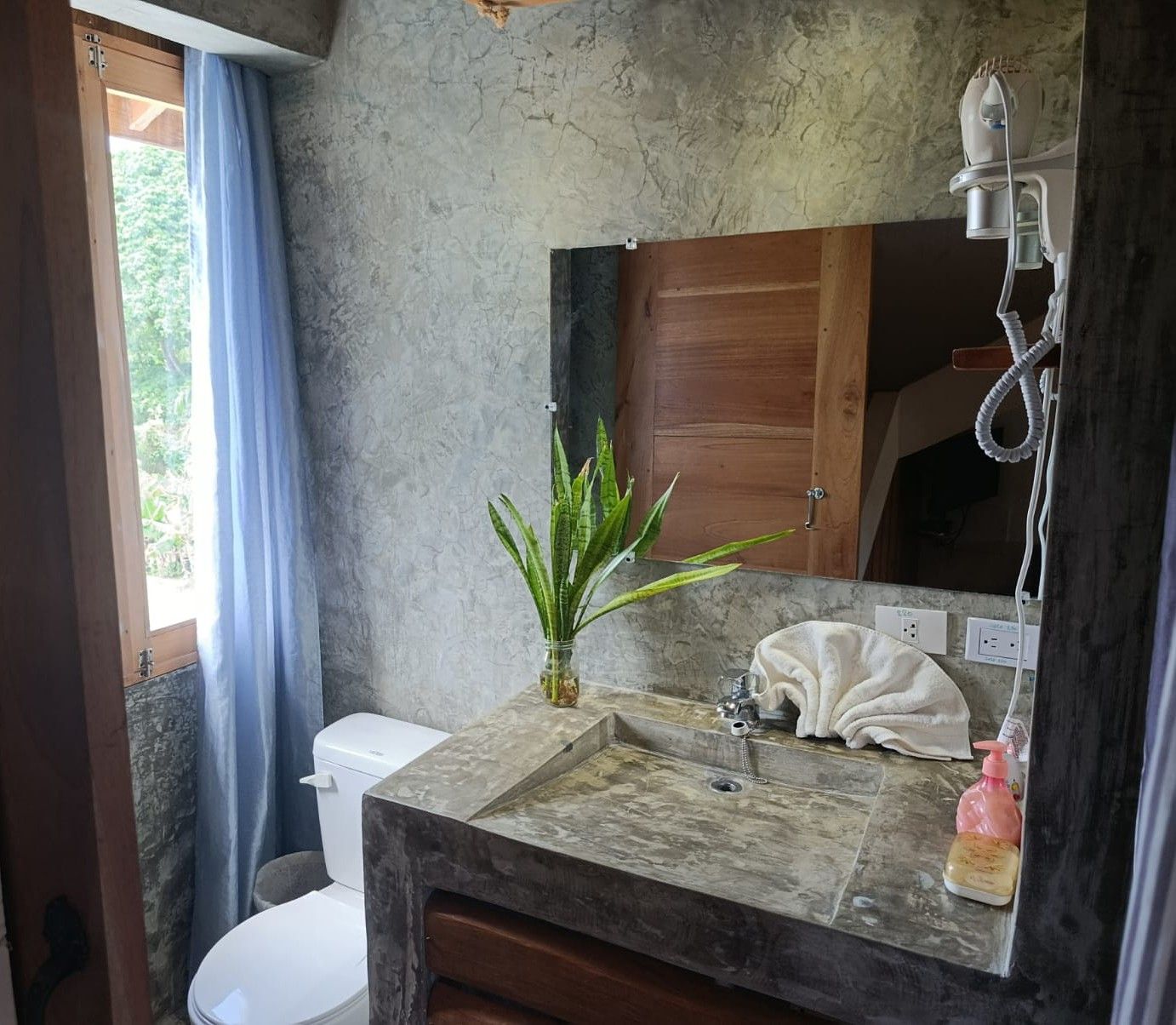 Bathroom with concrete sink, wooden mirror frame, toilet, blue curtain, and plants.