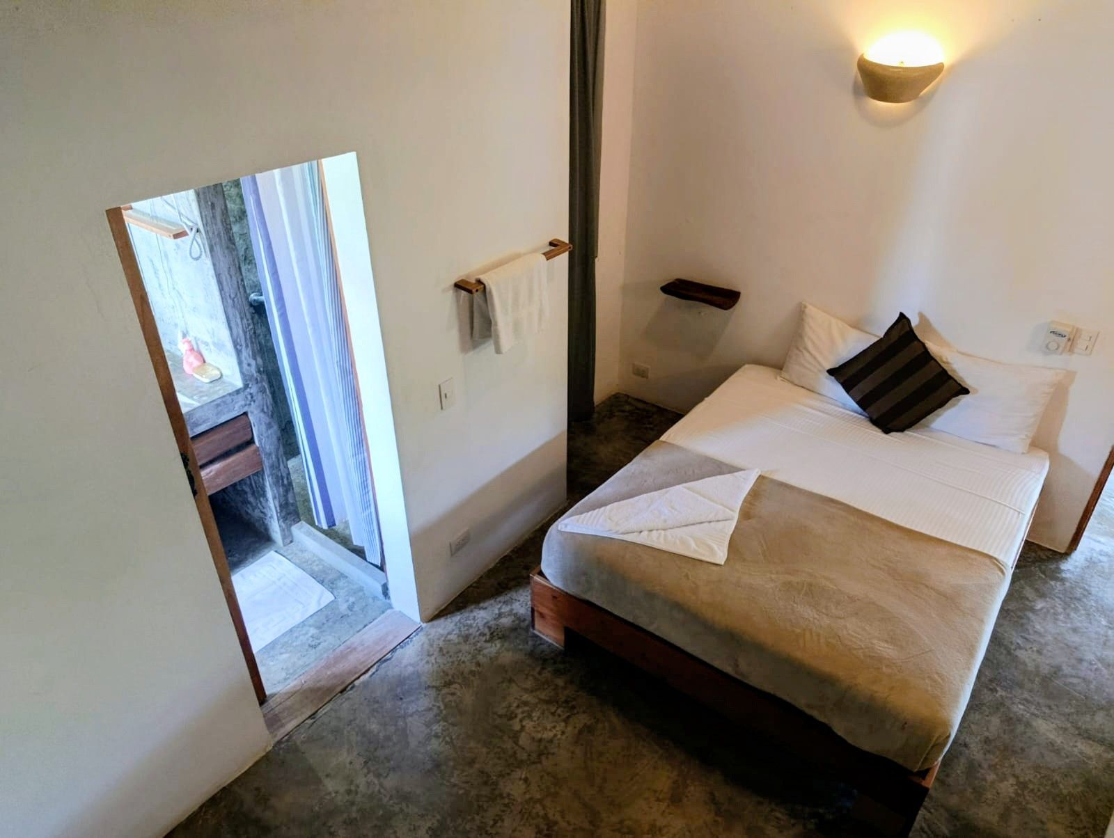 Bedroom with bed, towel rack, open doorway to bathroom. Concrete floor and white walls.
