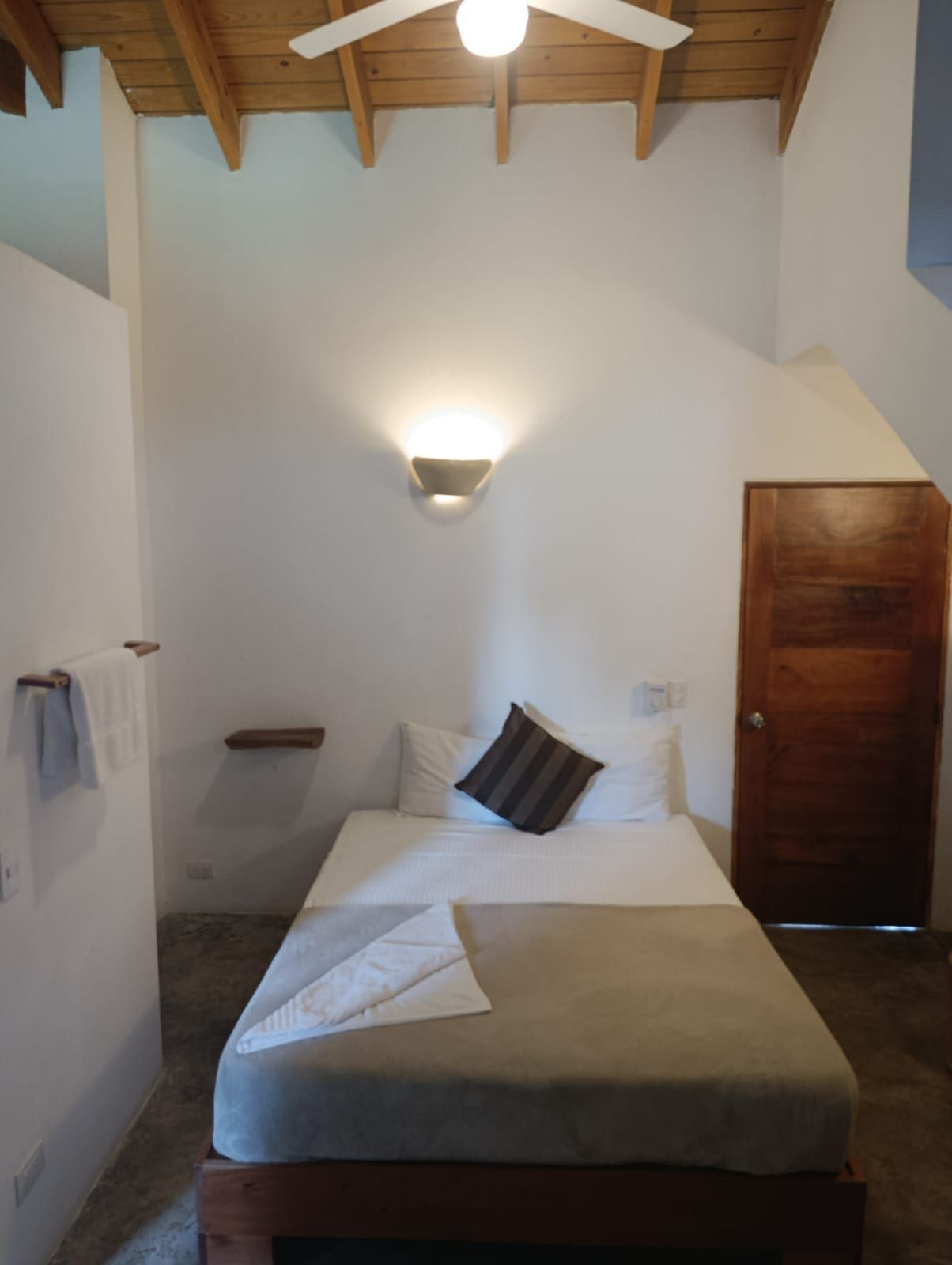 Small, white bedroom with a bed, wood door, and a wall-mounted light.