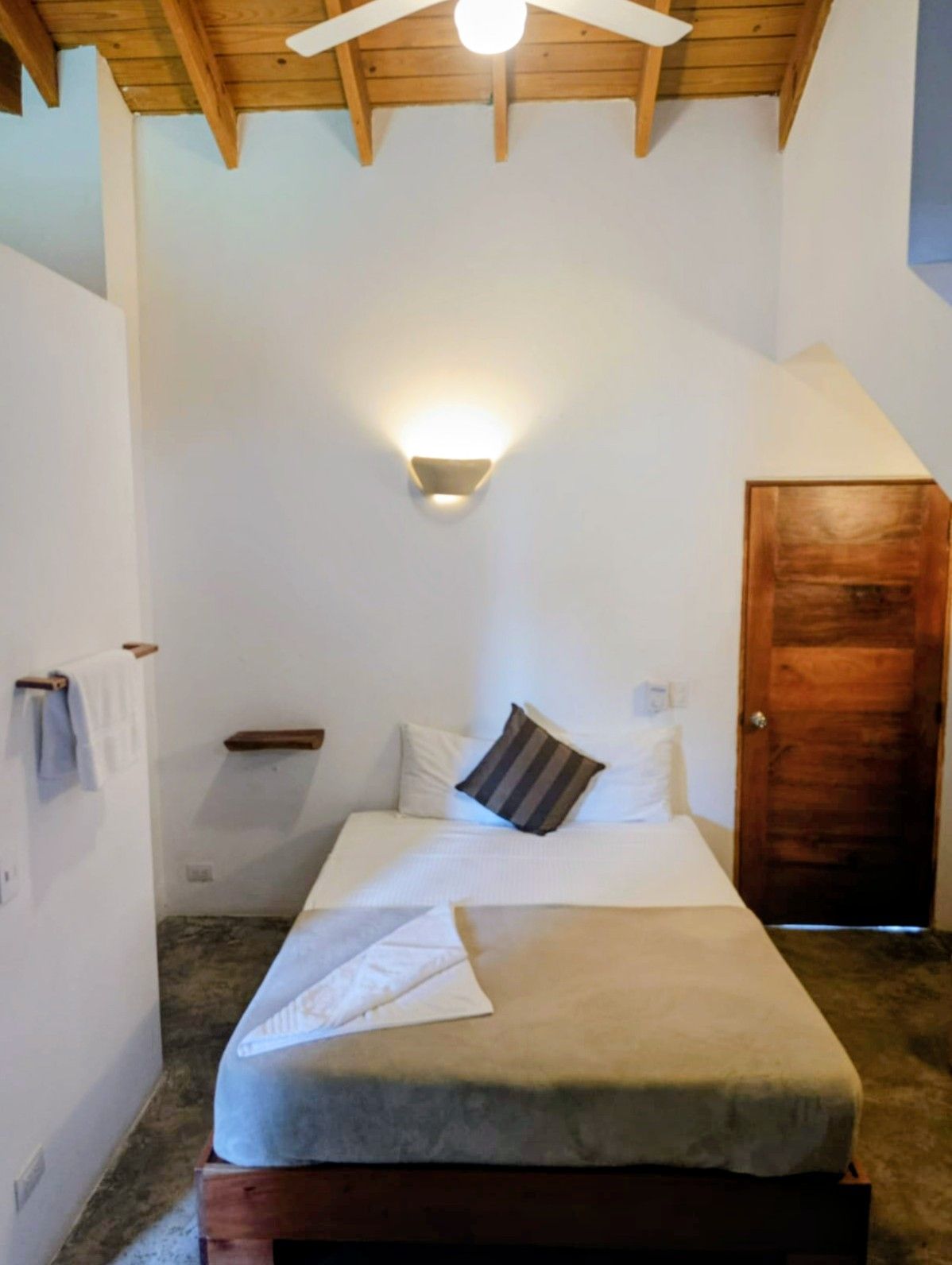 Small bedroom with a bed, a towel rack, and a closed wooden door. White walls, wood ceiling and grey floor.