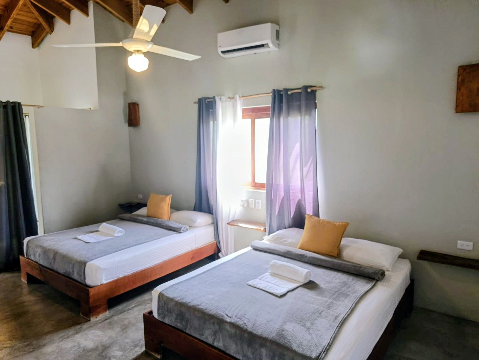 Bedroom with two beds, light gray walls, window with blue and purple curtains, ceiling fan.