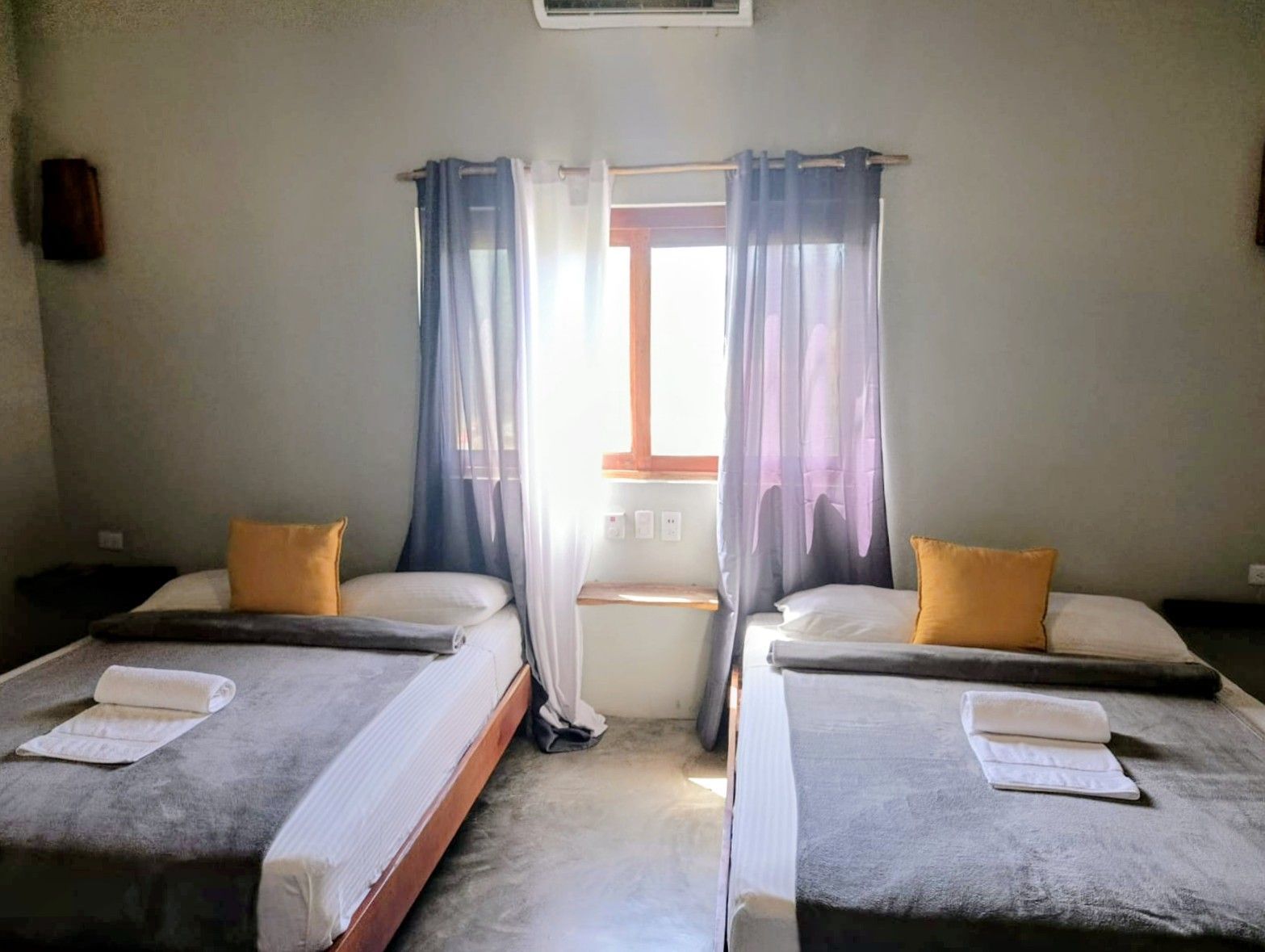 Two beds in a room, window with curtains, gray walls, yellow pillows, towels.