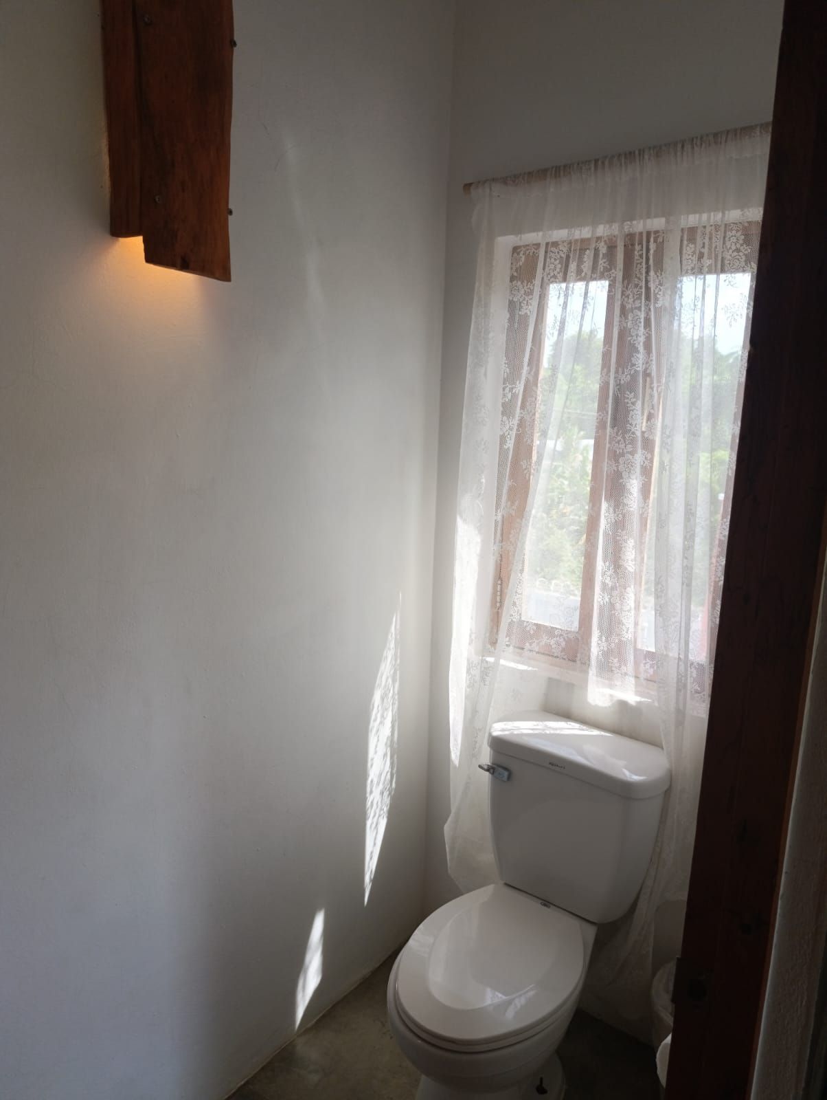 Small white bathroom with a toilet by a window with lace curtains. A wooden light fixture is on the wall.
