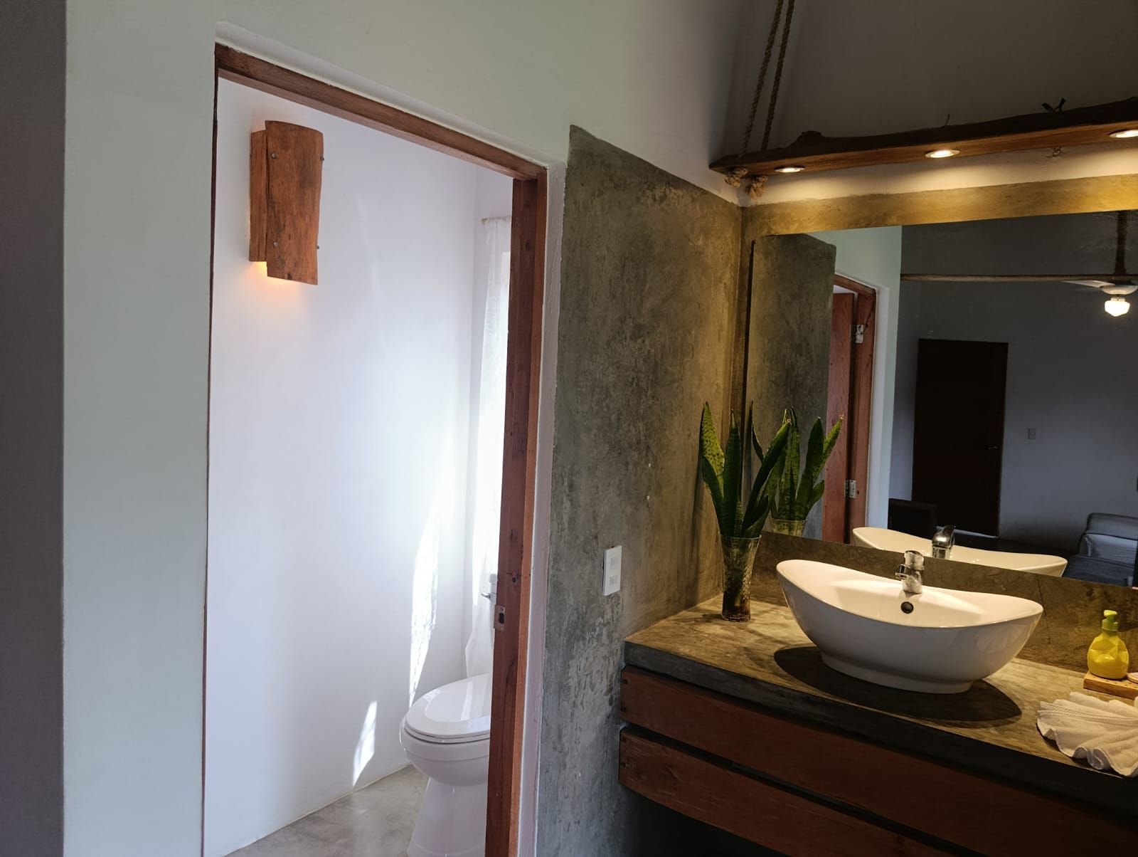 Bathroom with toilet in white-walled room, vessel sink on wooden vanity, wooden sconce, and textured wall.