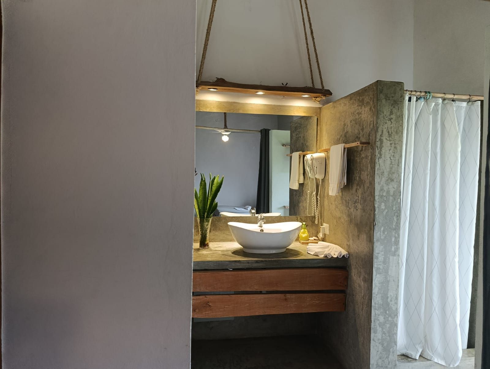 Bathroom with a concrete wall and wooden vanity, white sink, and shower curtain; mirror reflection.