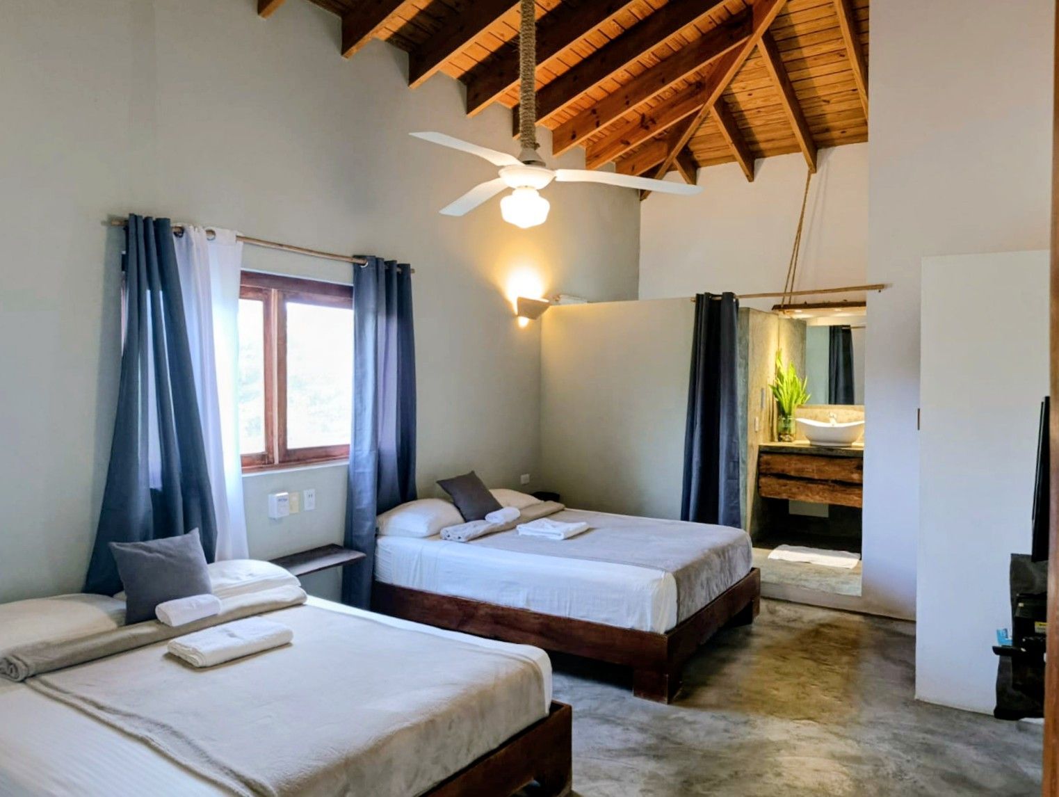 Bedroom with two beds, high ceiling with fan, windows with blue curtains, and a partially visible bathroom.