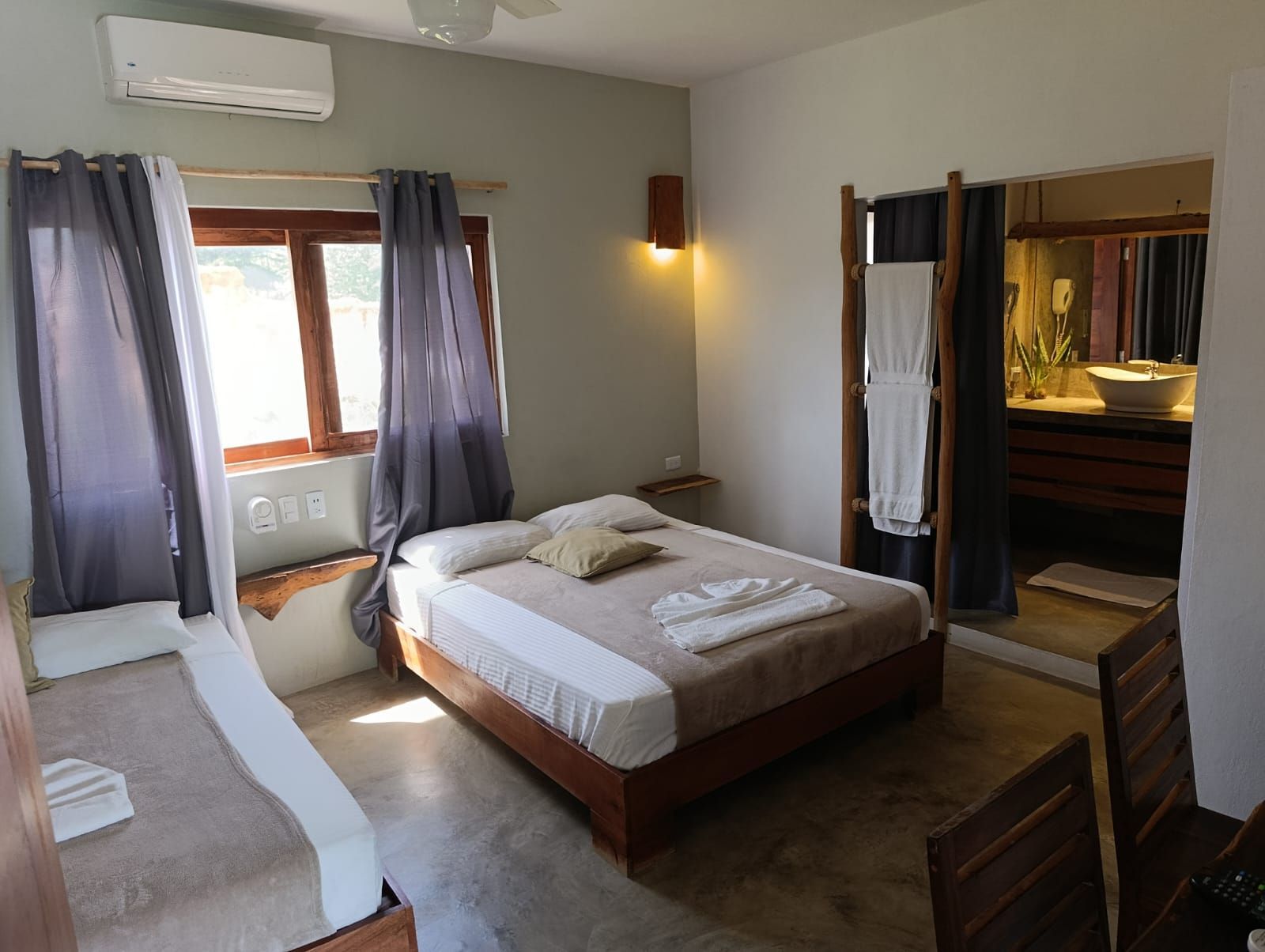 Bedroom with two beds, window, and open bathroom. Grey curtains, beige linens, and wooden furniture.