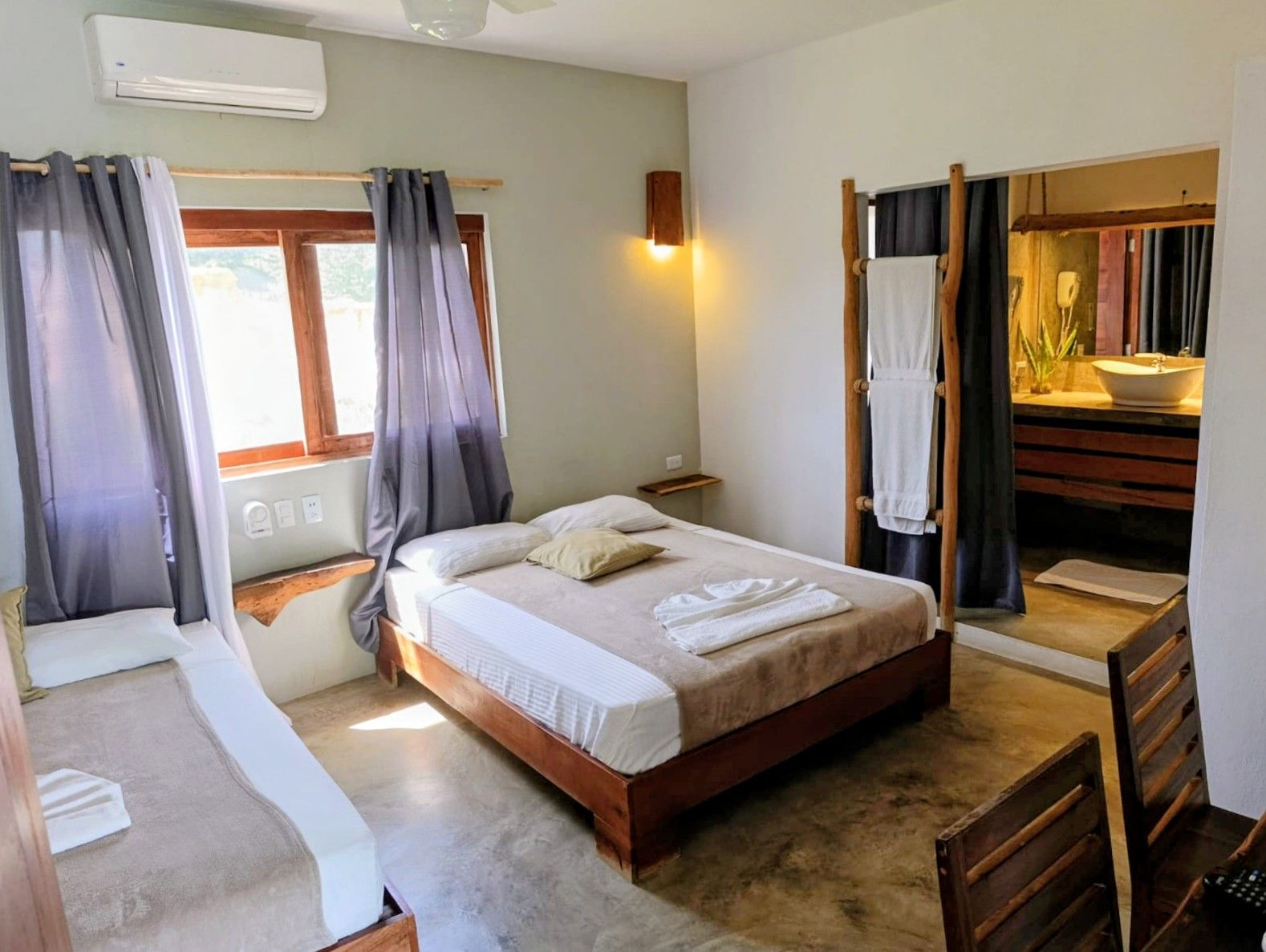 Bedroom with two beds, window, and open bathroom. Grey curtains, wooden furniture, and light-colored bedding.