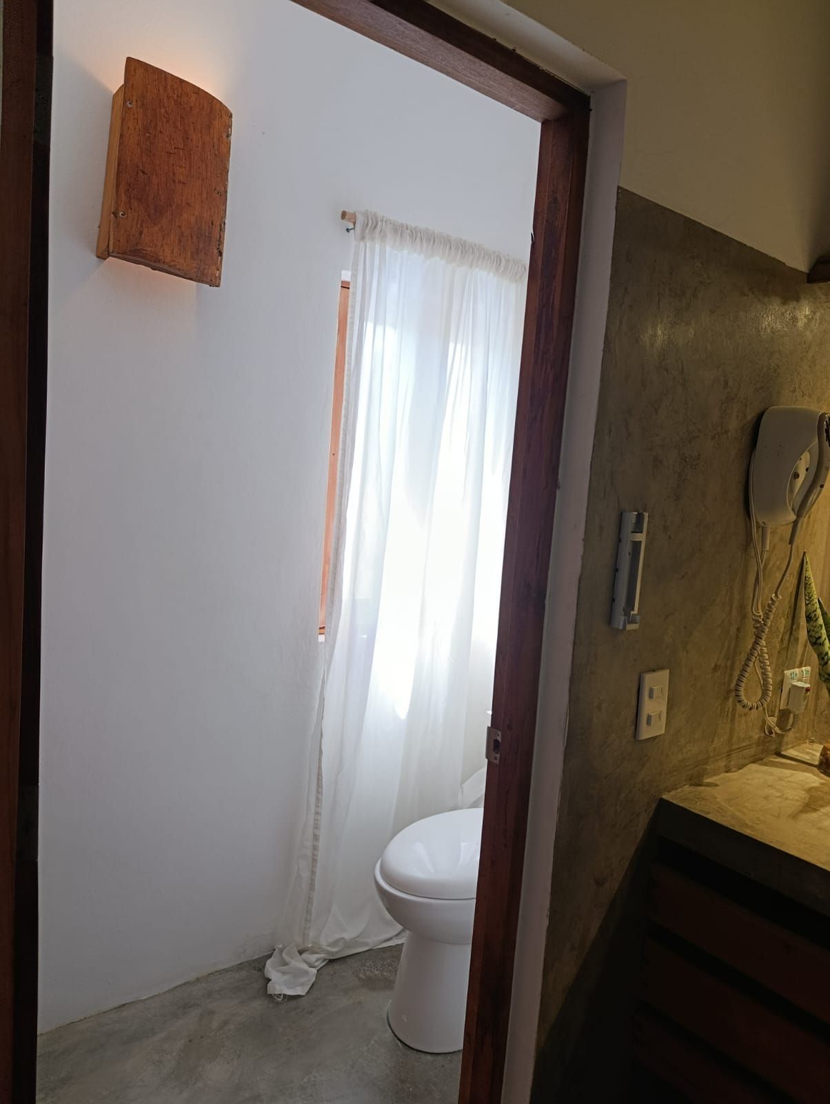 View into a bathroom with a toilet, window with sheer curtain, and an orange wall sconce.
