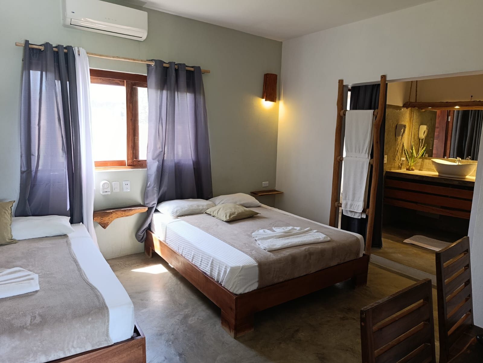 Bedroom with two beds, window, and open bathroom. Grey and tan color scheme.