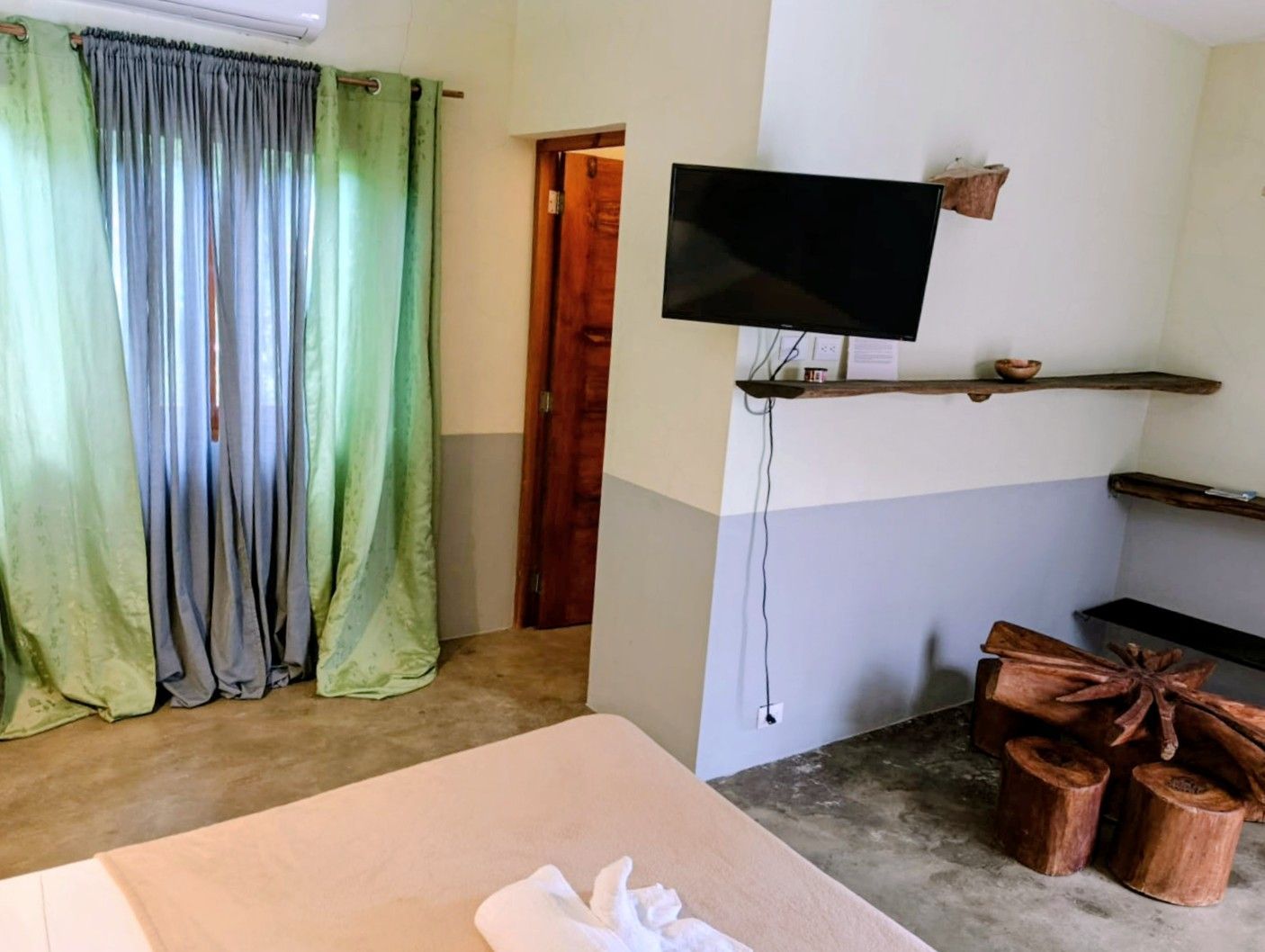 Bedroom with bed, TV, wooden stools, gray and green curtains, and a wooden door.