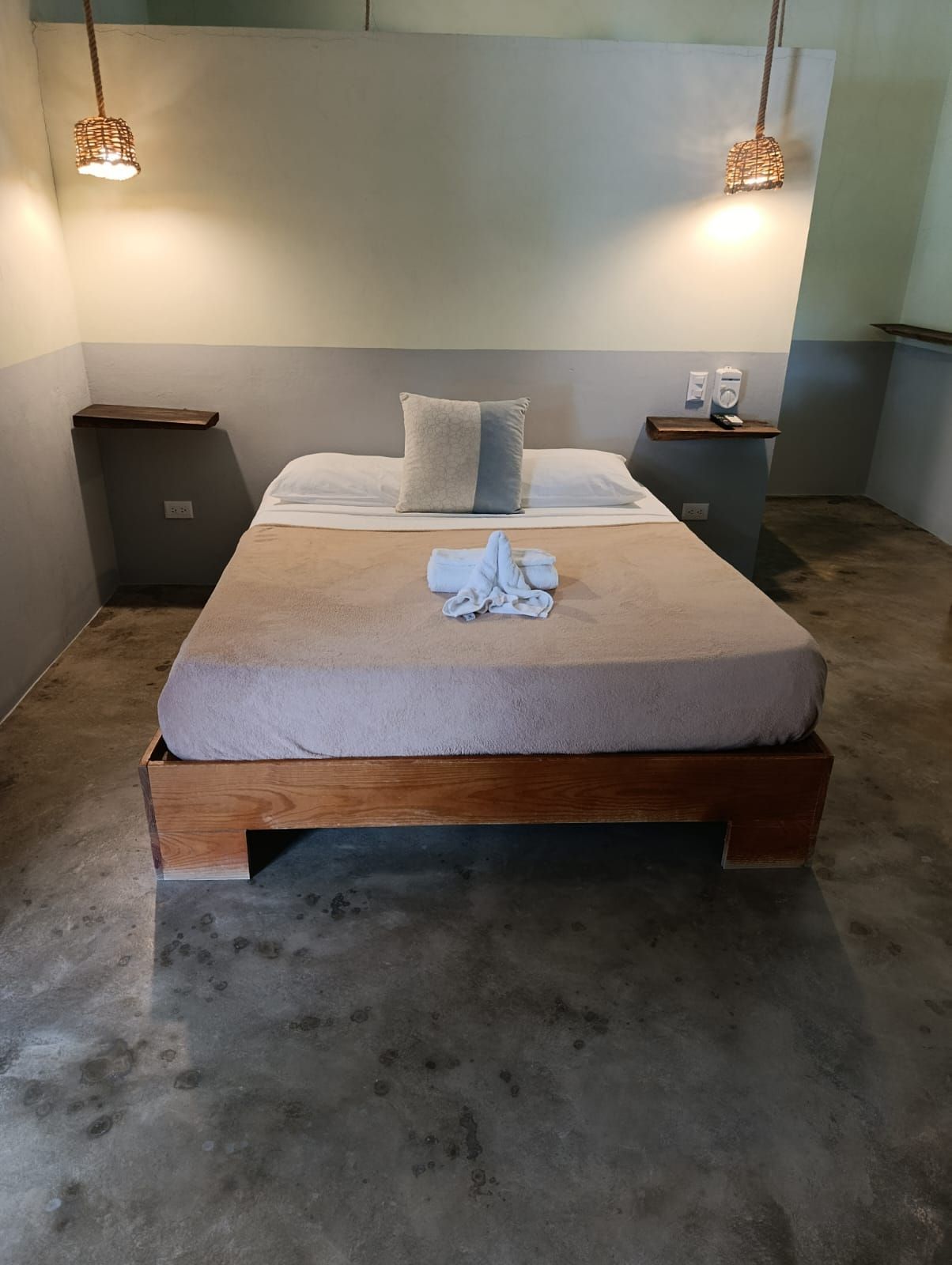 Bedroom with wooden bed frame, bedside tables, neutral walls, concrete floor, and hanging lights.
