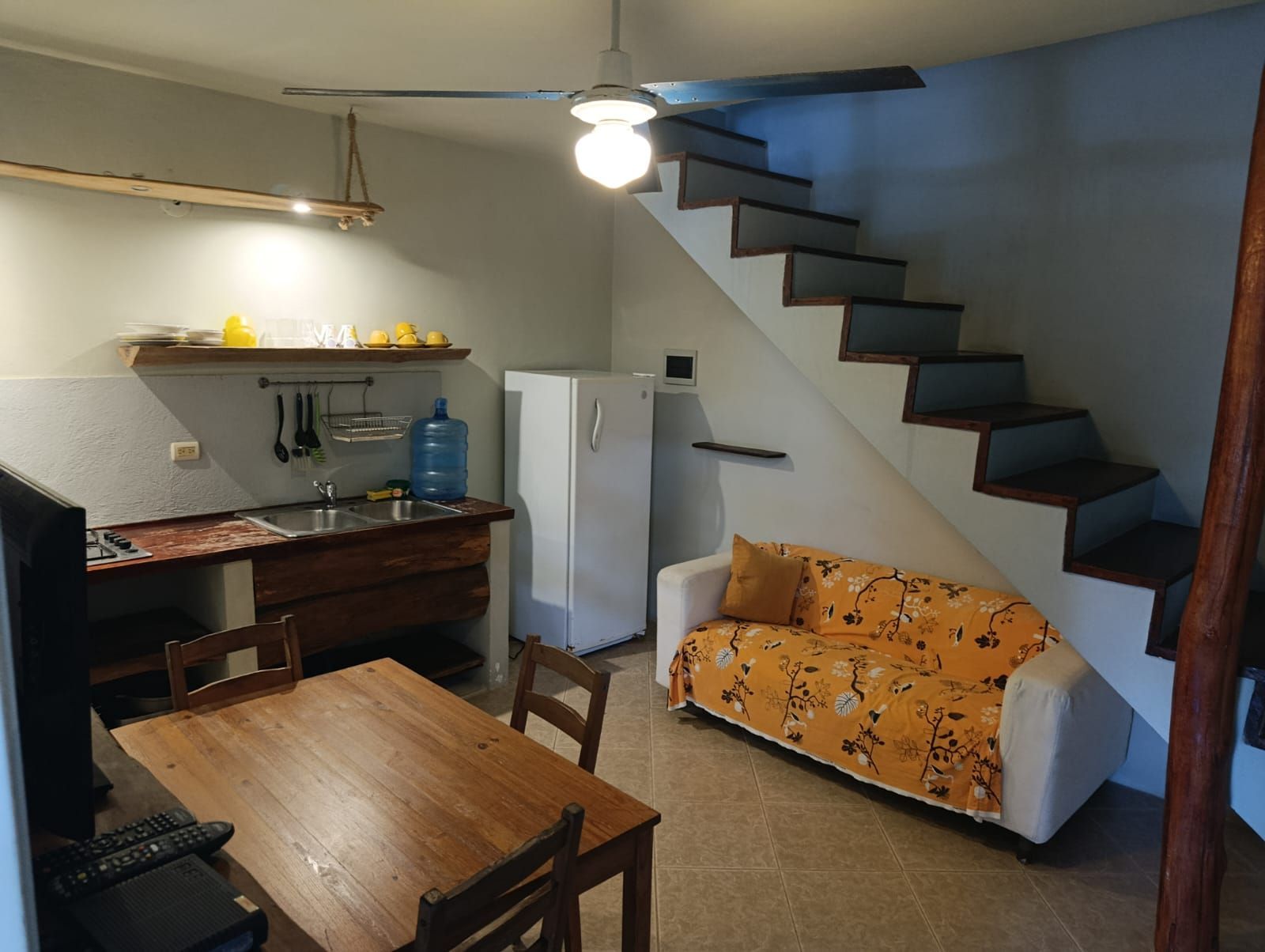 Small kitchen/living area with stairs. Wooden table, sofa with orange cover, fridge, and overhead light.